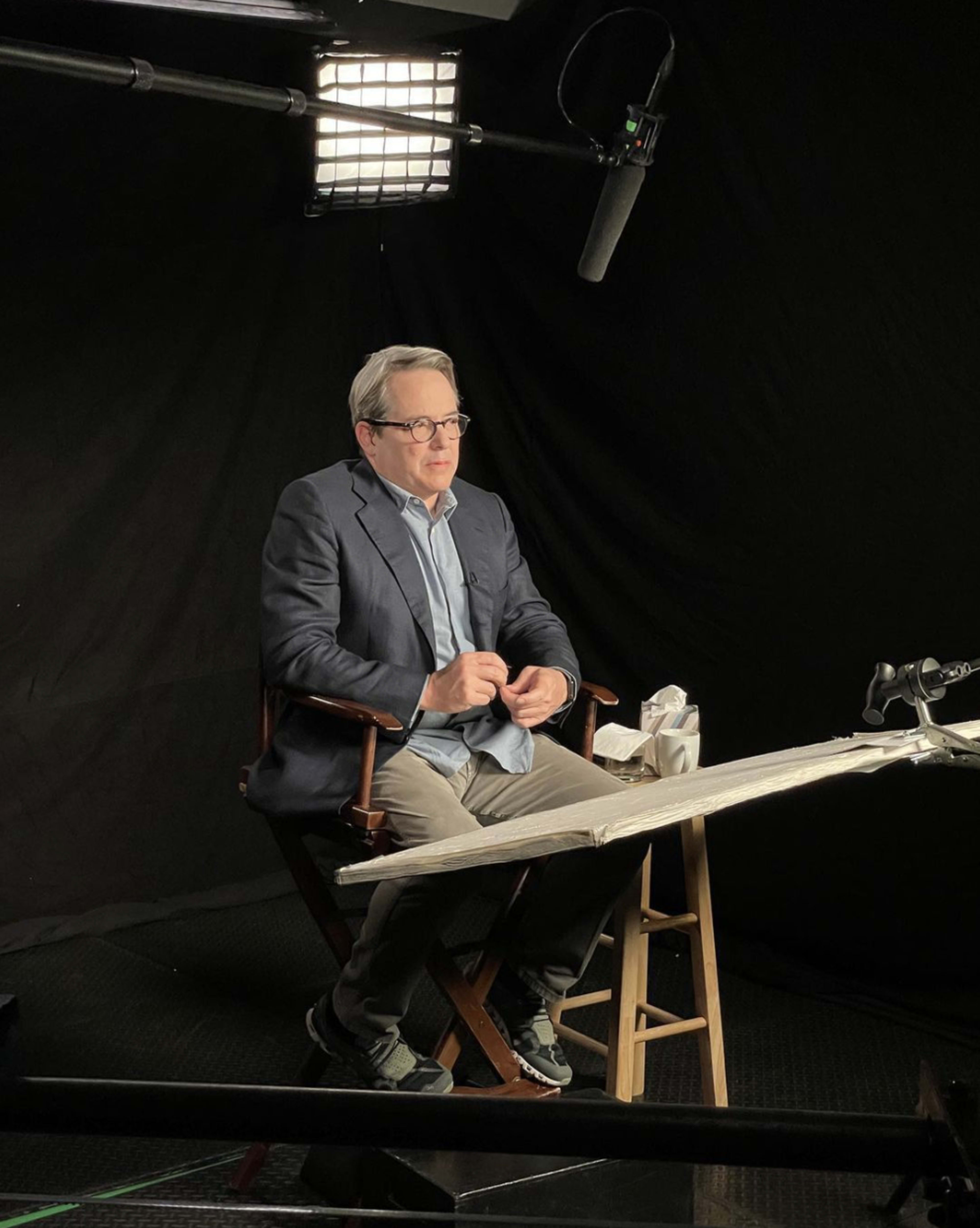 A man is seated in a director's chair in front of a black backdrop, with a microphone overhead and a table in front of him.