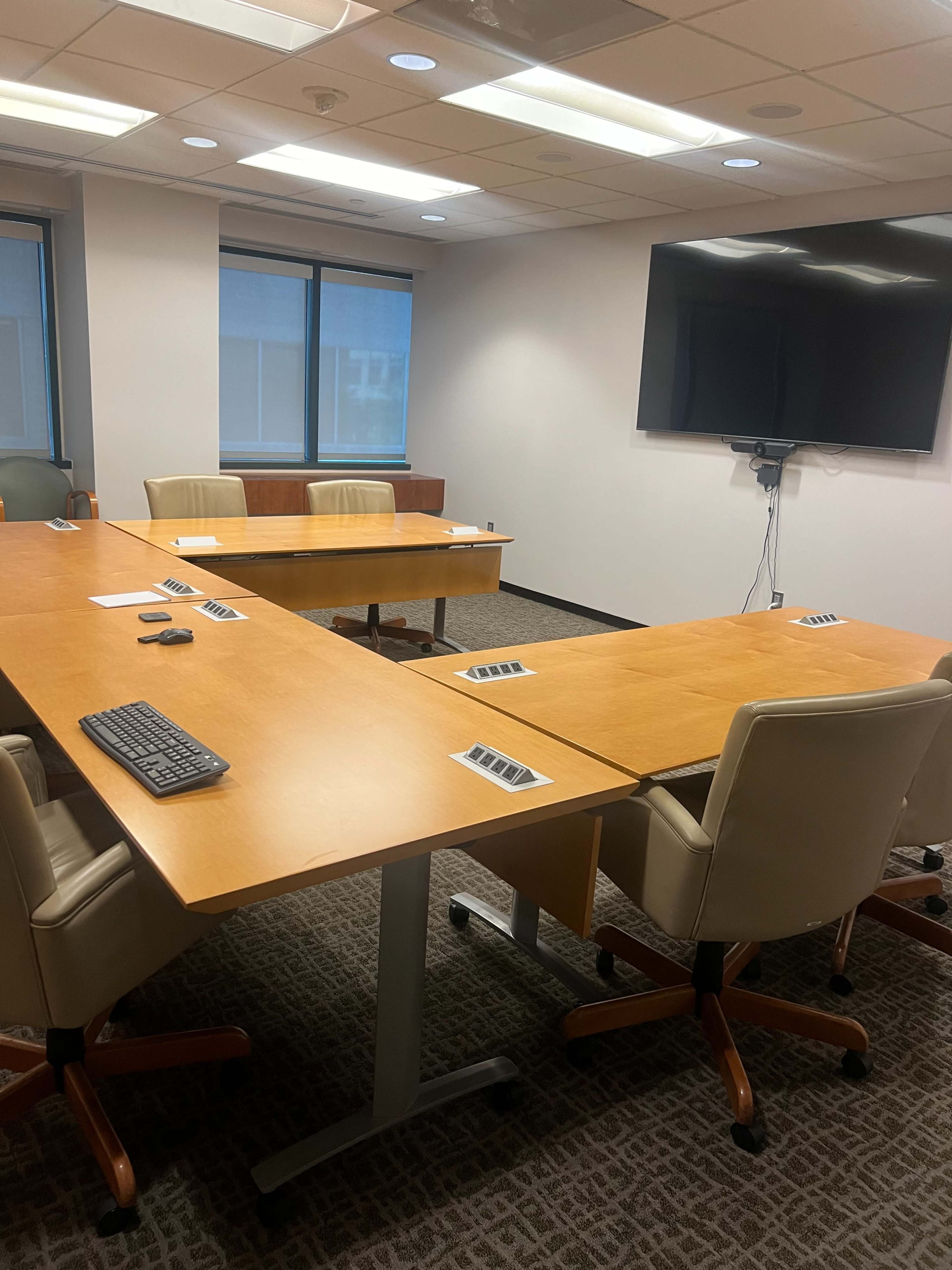 A modern conference room features two wooden tables arranged in a U-shape, with several beige chairs and a large wall-mounted screen.