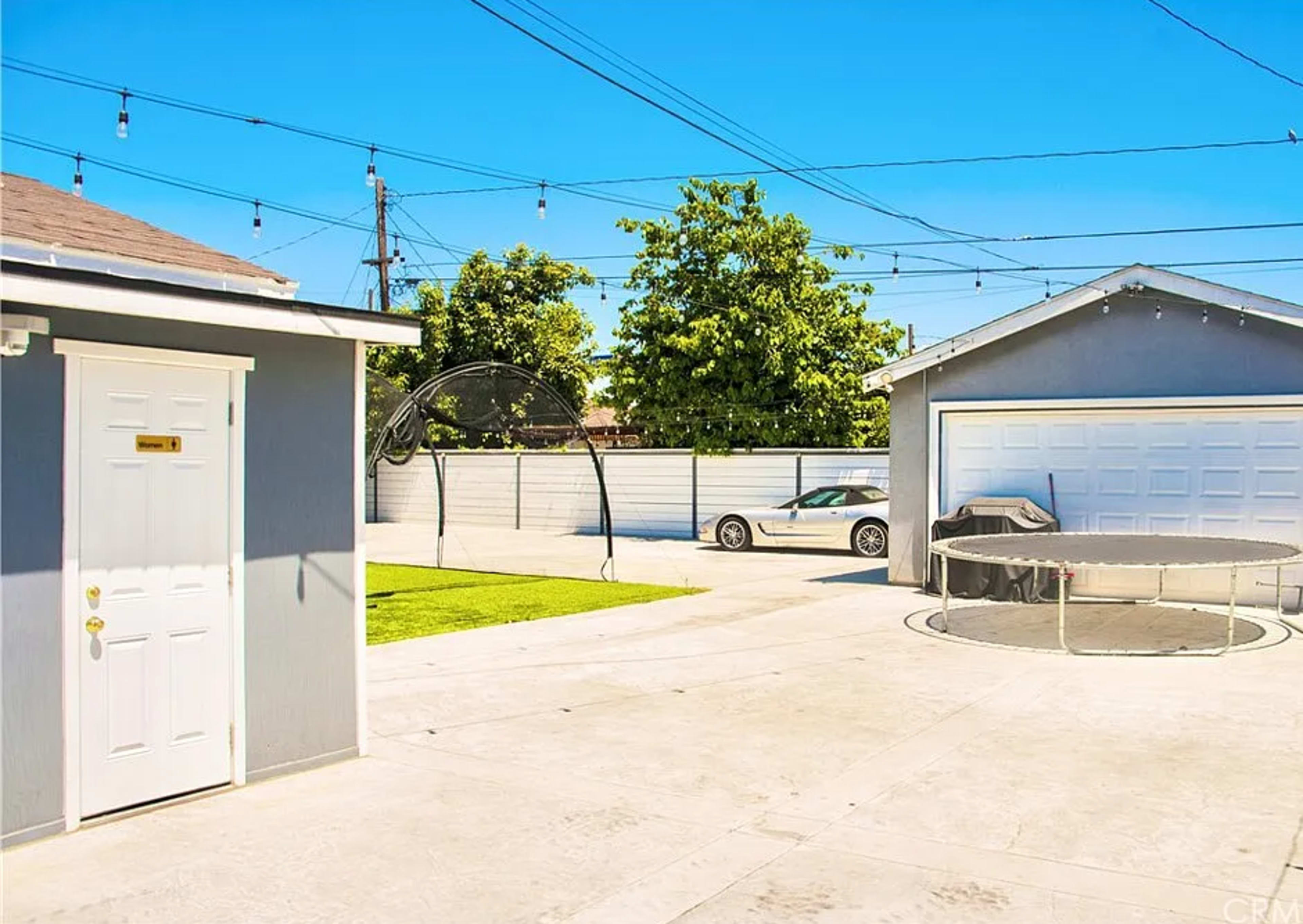The image shows a backyard with a gray shed, a trampoline, a parked car, and overhead string lights under a clear blue sky.