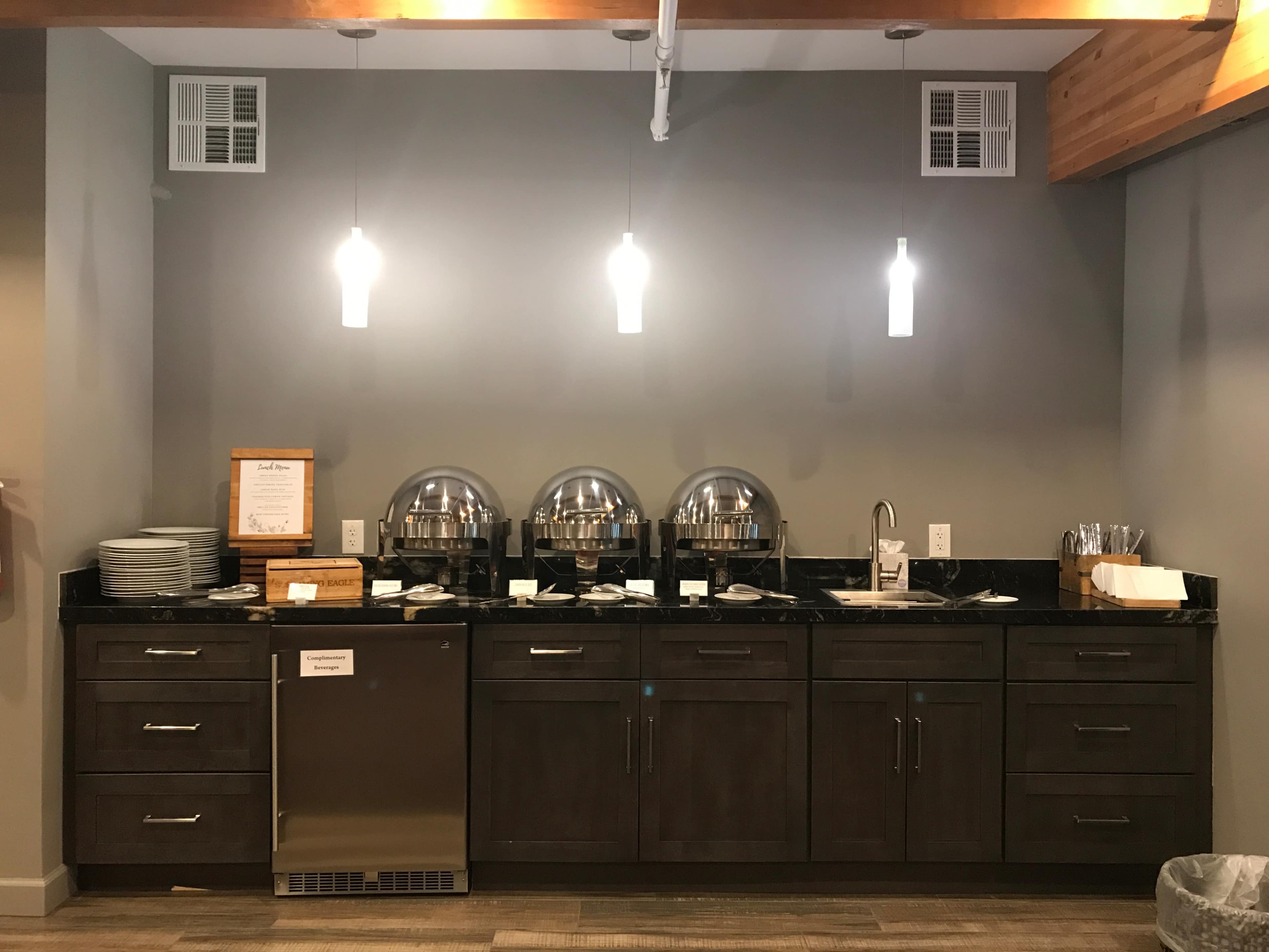 A modern buffet station with a dark countertop, several chafing dishes, plates, utensils, and a stainless-steel refrigerator against a gray wall.