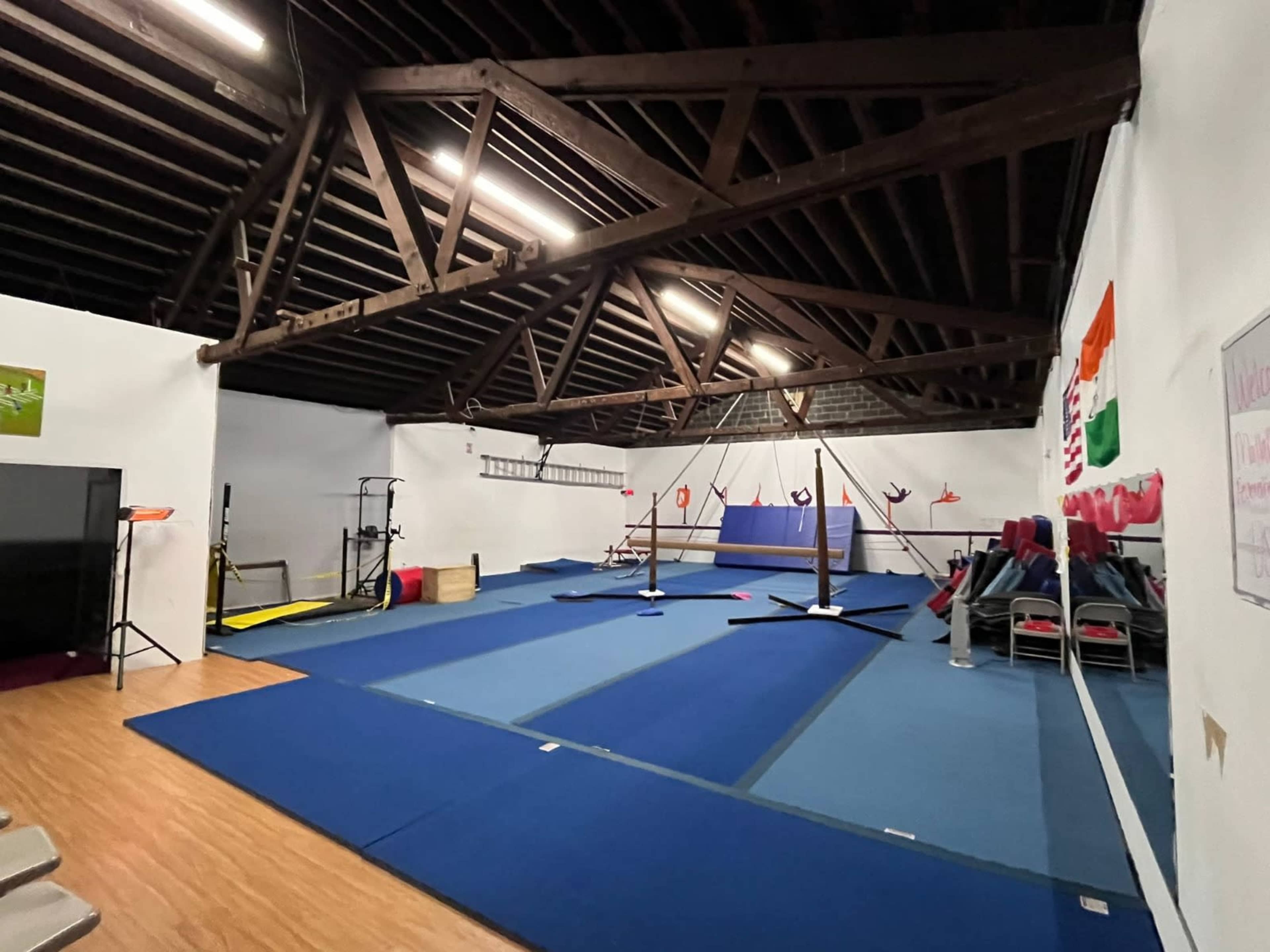 A spacious gymnasium with wooden beams, featuring gymnastics equipment and blue mats spread across the floor.