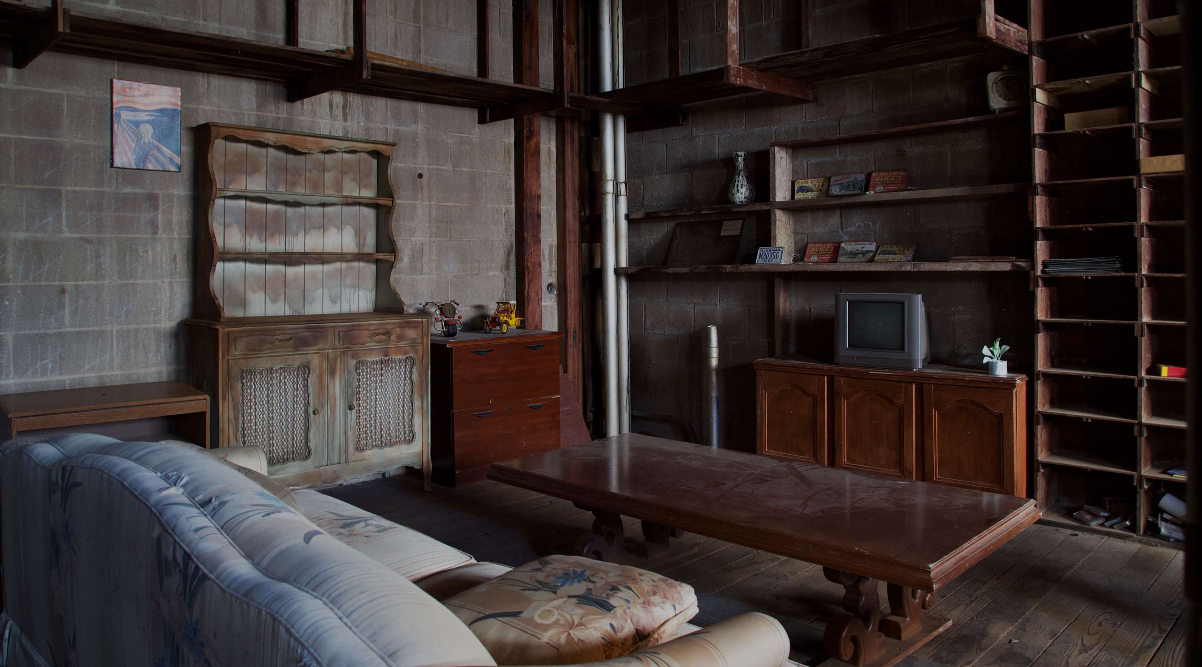 A living room features a wooden coffee table, a couch, and a set of shelves against a concrete wall, with an old television and handcrafted furniture.
