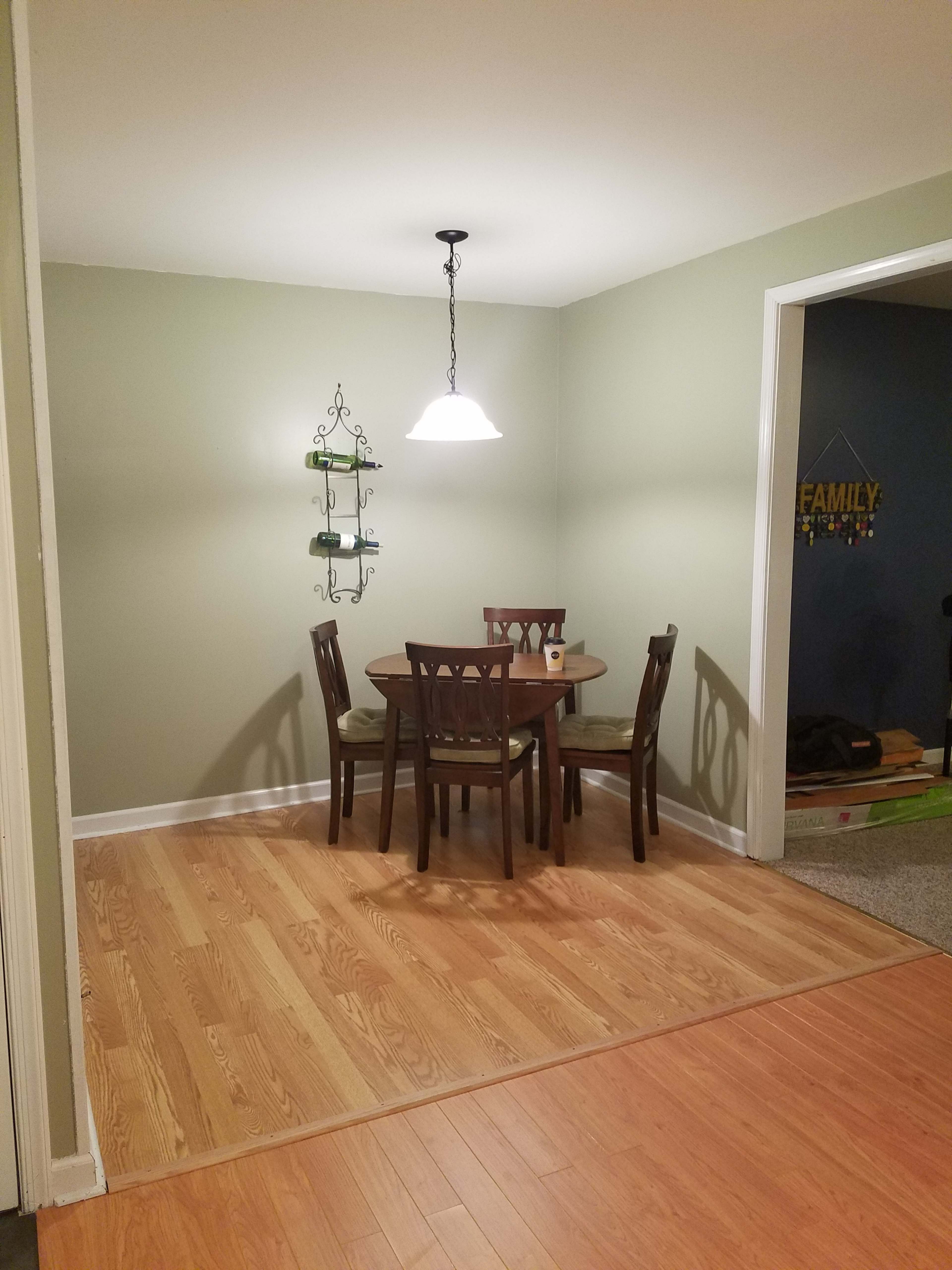 A small dining area features a wooden table with four chairs, illuminated by a pendant light, and a green wall with a decorative shelf.
