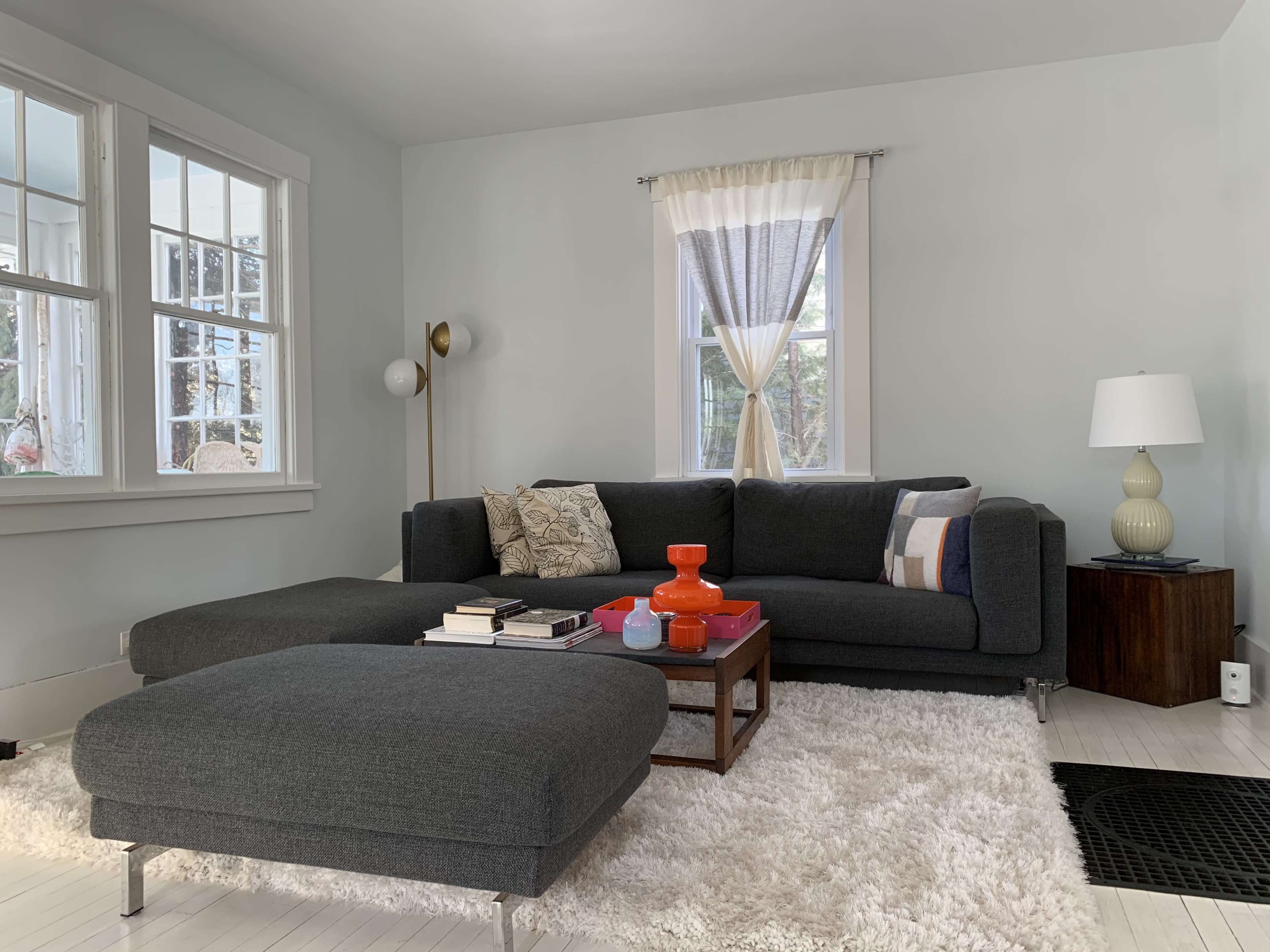 The living room features a dark gray sectional couch, a light-colored rug, a small wooden table, and large windows allowing natural light to enter.