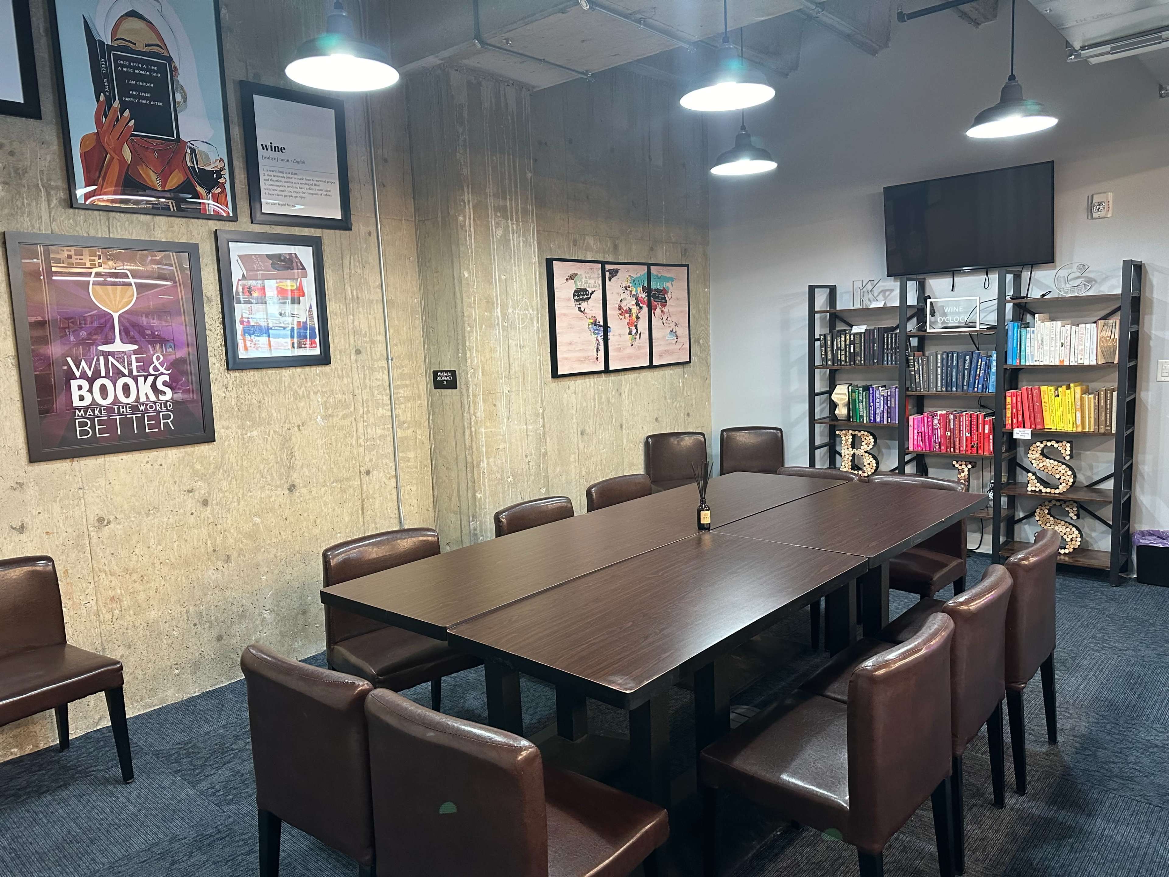 A conference room with a large table surrounded by brown chairs, featuring colorful artwork and shelves filled with books against a concrete wall.