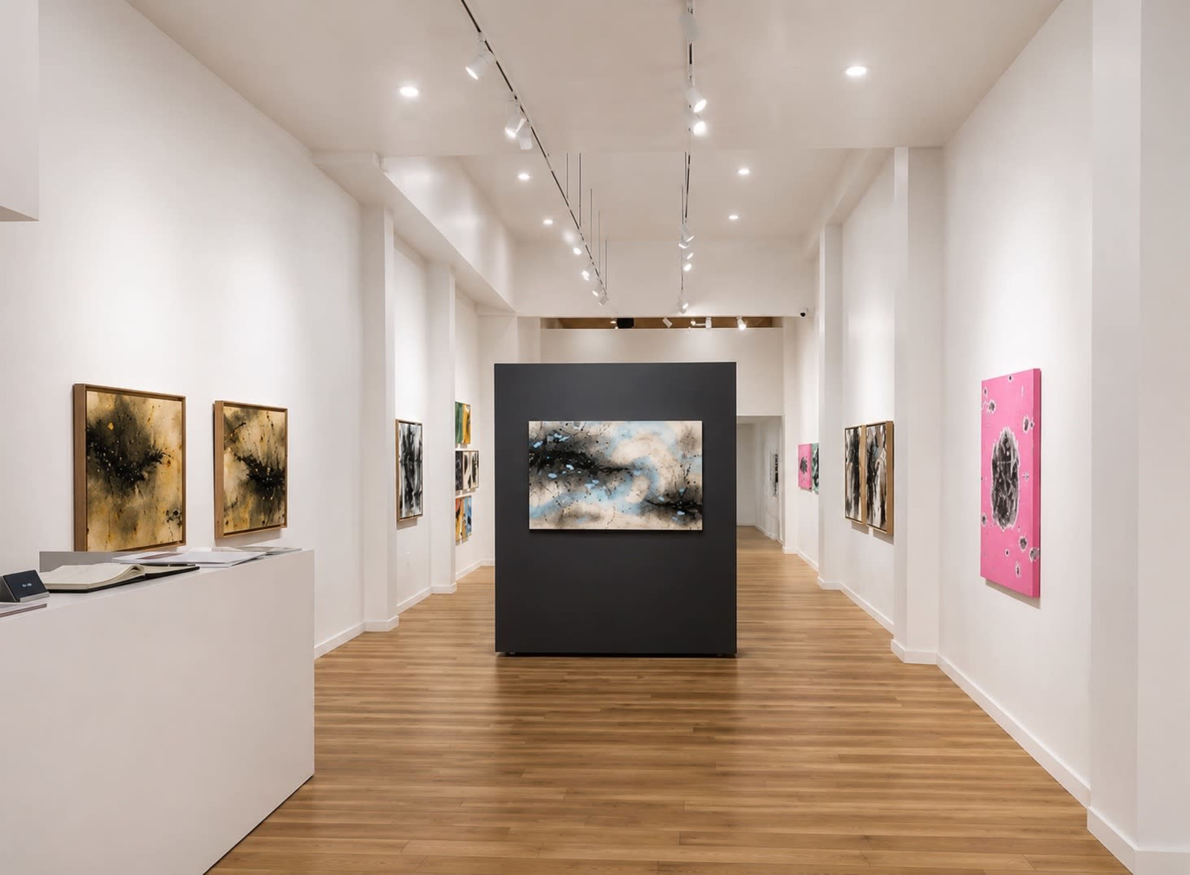 The image shows an art gallery with paintings displayed on white walls, featuring a central dark display panel showcasing a large artwork, while warm wooden floors extend throughout the space.