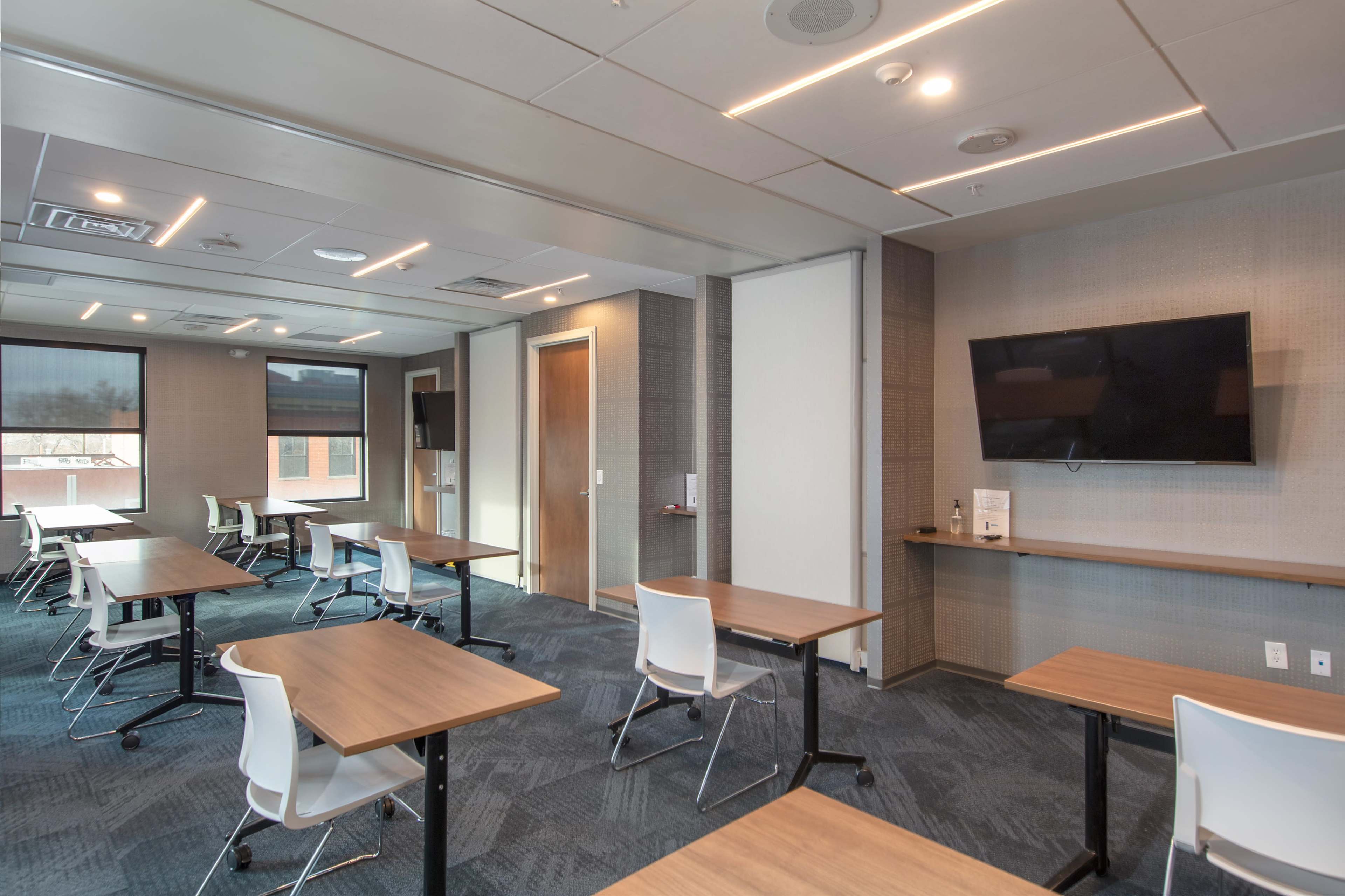 A modern classroom with several tables and chairs arranged for instruction, featuring a wall-mounted television and large windows.