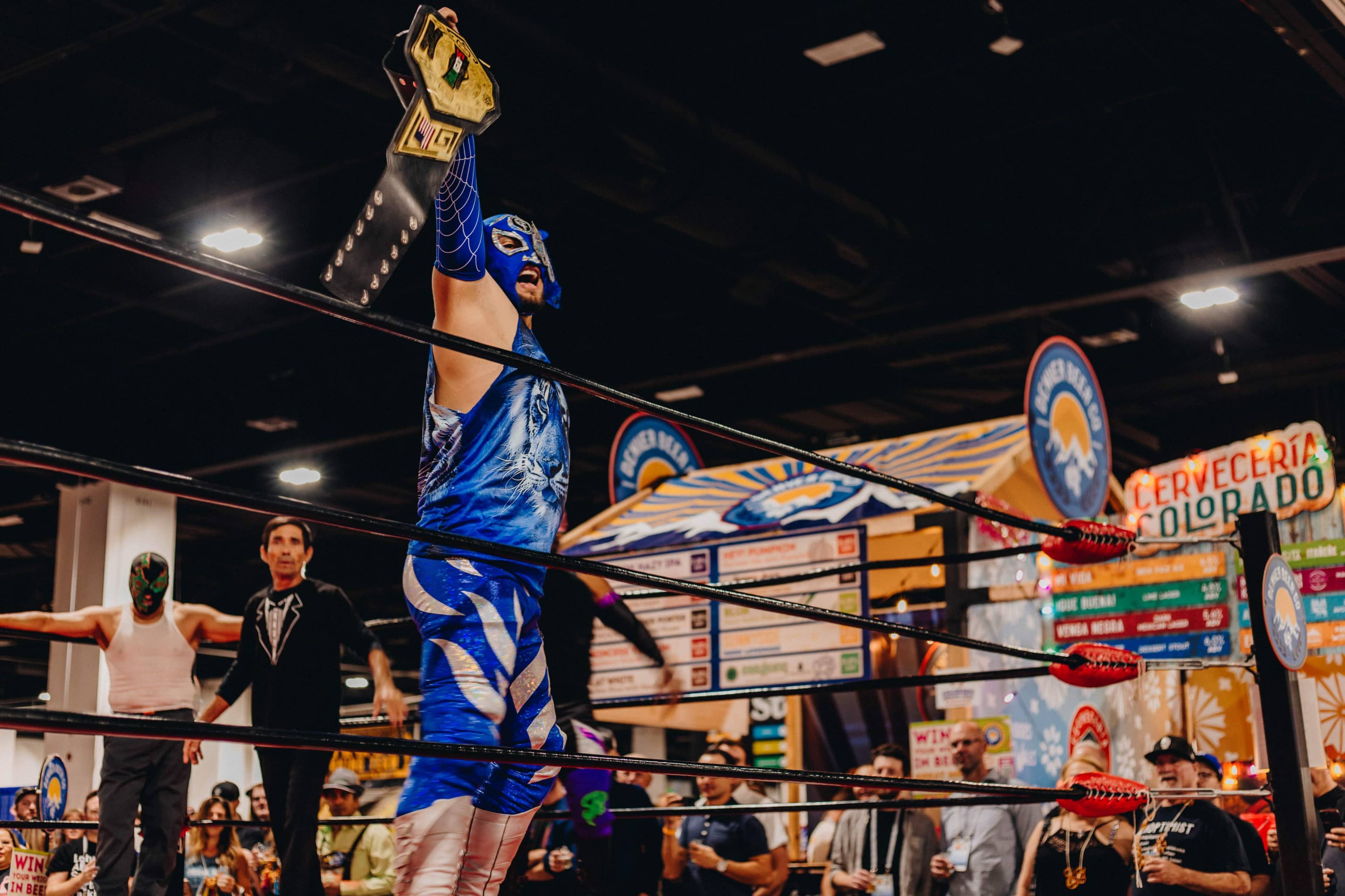 A masked wrestler in a blue costume raises a championship belt in a wrestling ring surrounded by an audience and event signage.