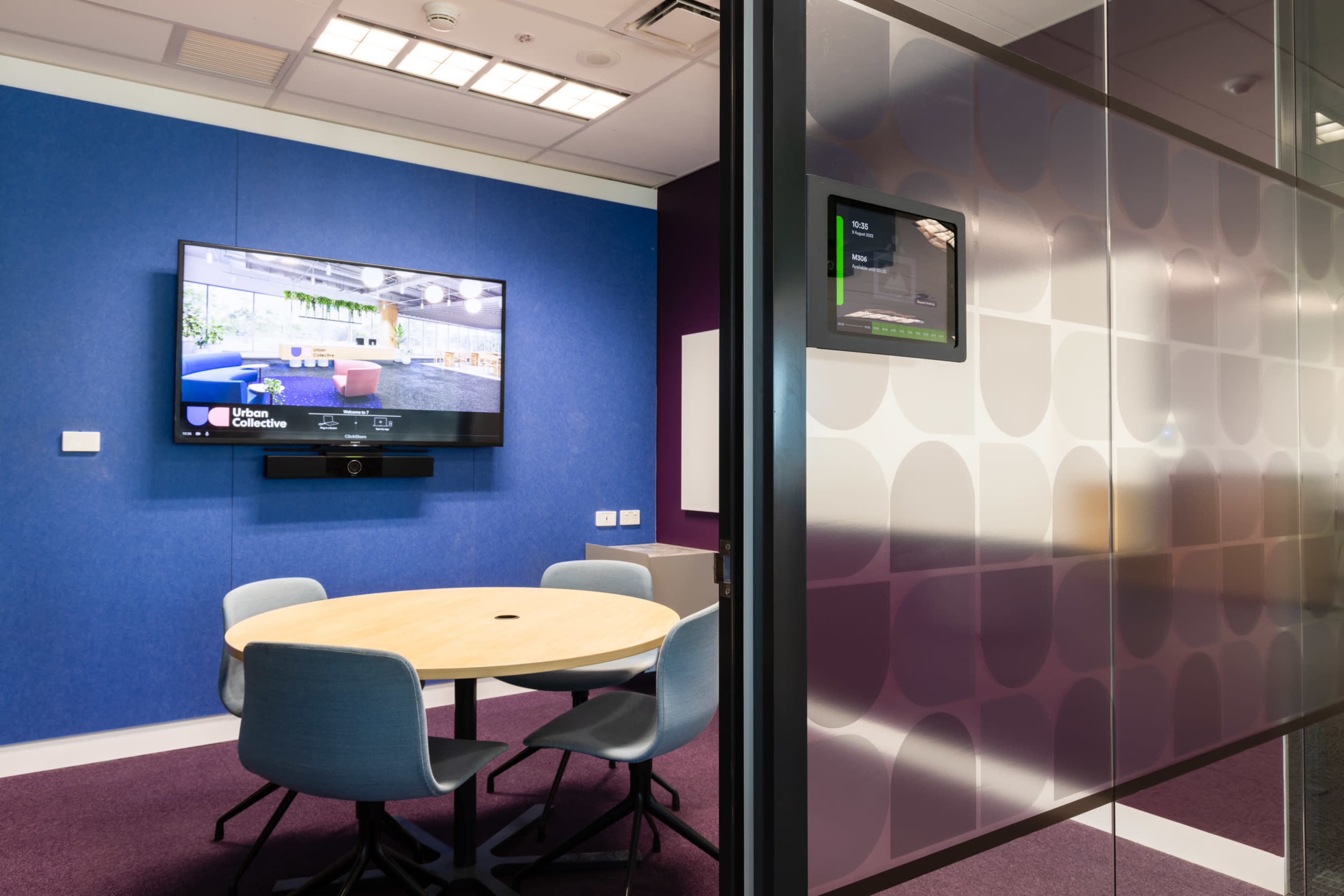 A conference room features a circular table surrounded by chairs, with a large screen displaying a video on the wall and a control panel mounted adjacent to a glass door.