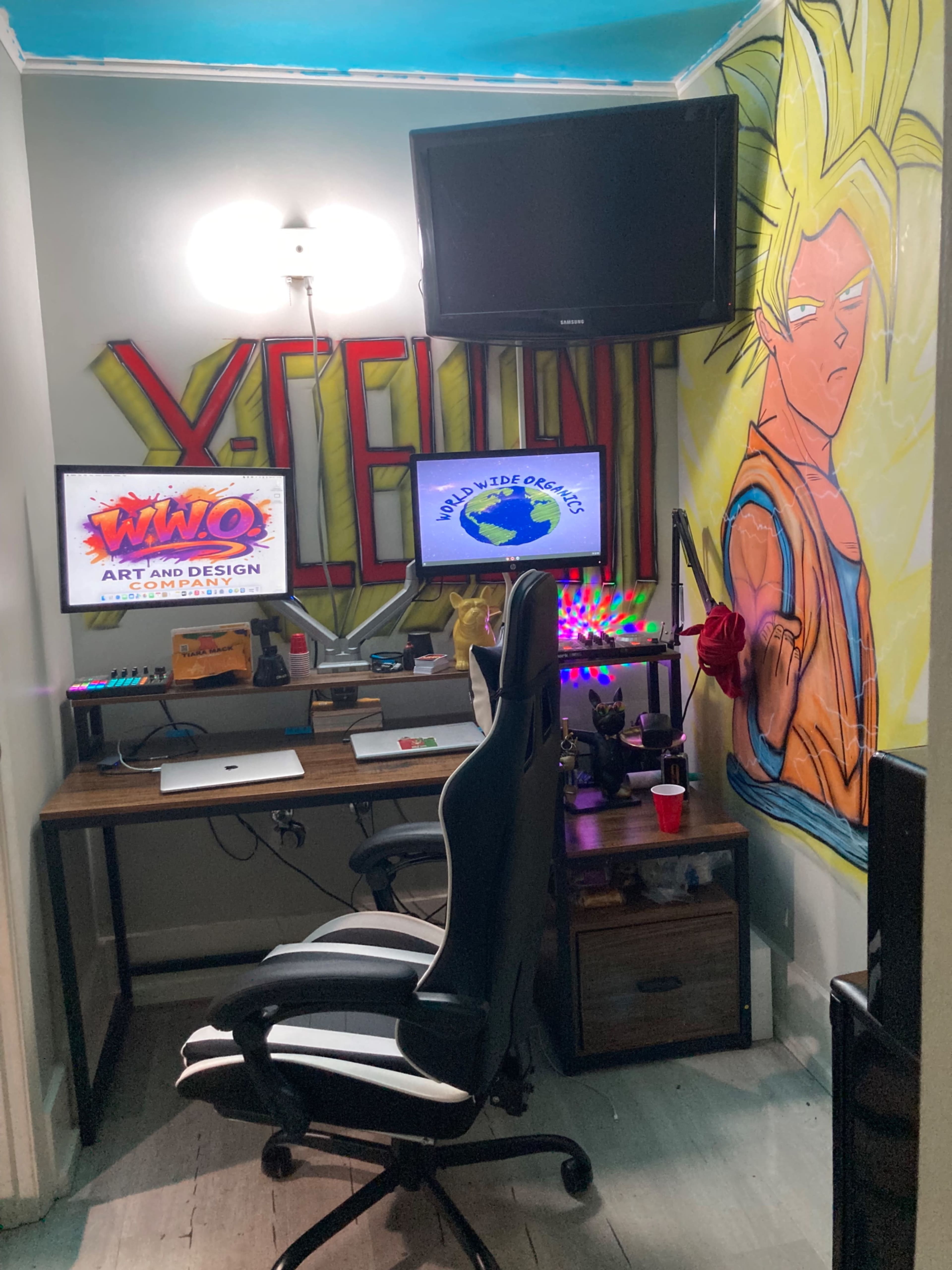 The image shows a small workspace with a desk set up, featuring two computer monitors, a gaming chair, and colorful wall art, including a large character illustration.