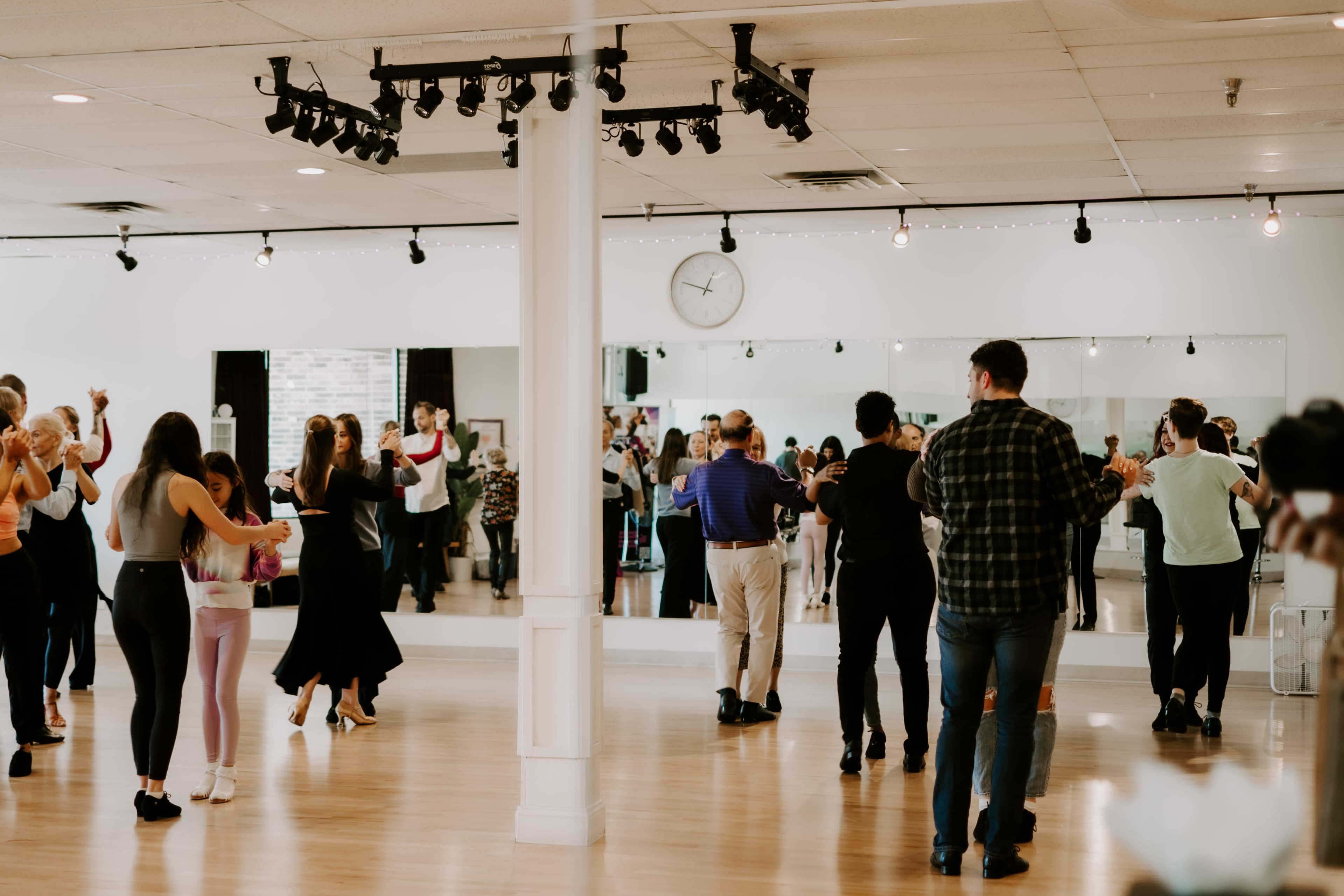 A dance studio features several couples and groups practicing various dance styles in front of large mirrors.