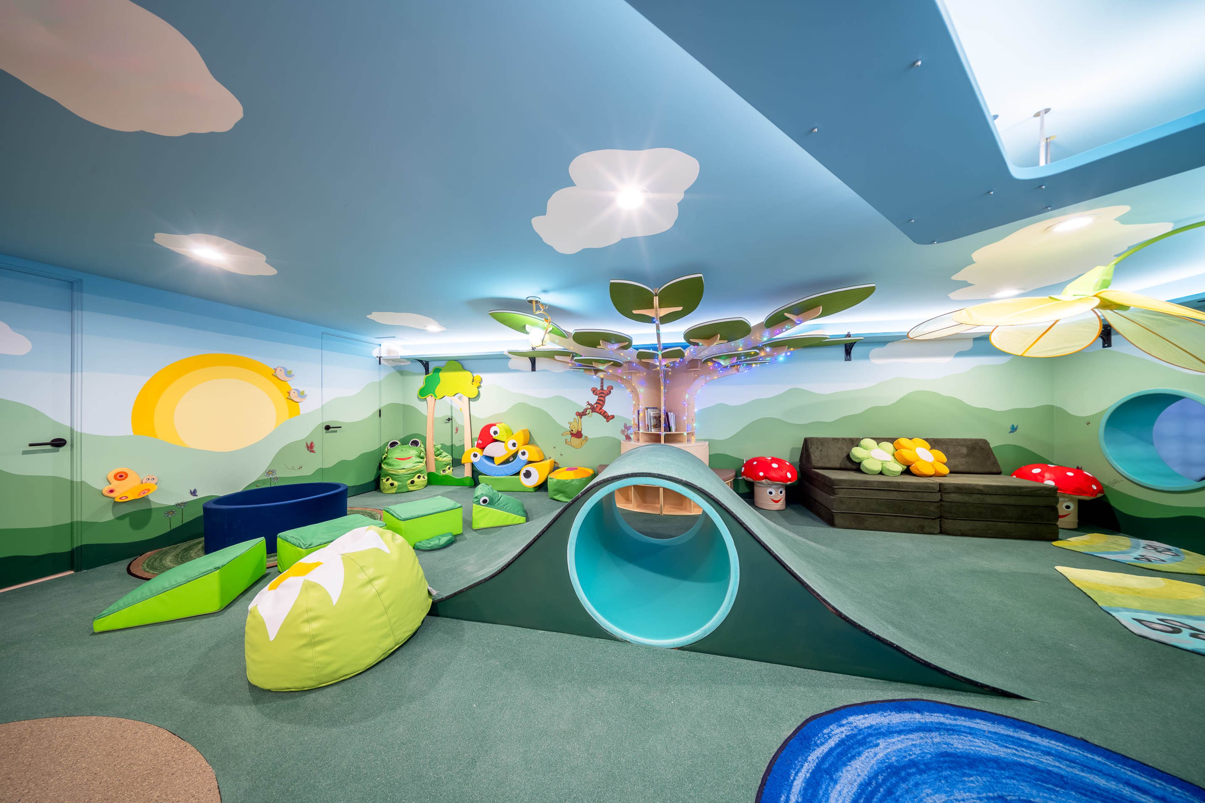 A colorful children's playroom features a slide, soft tunnels, and various playful decorations on the walls, including trees and clouds.