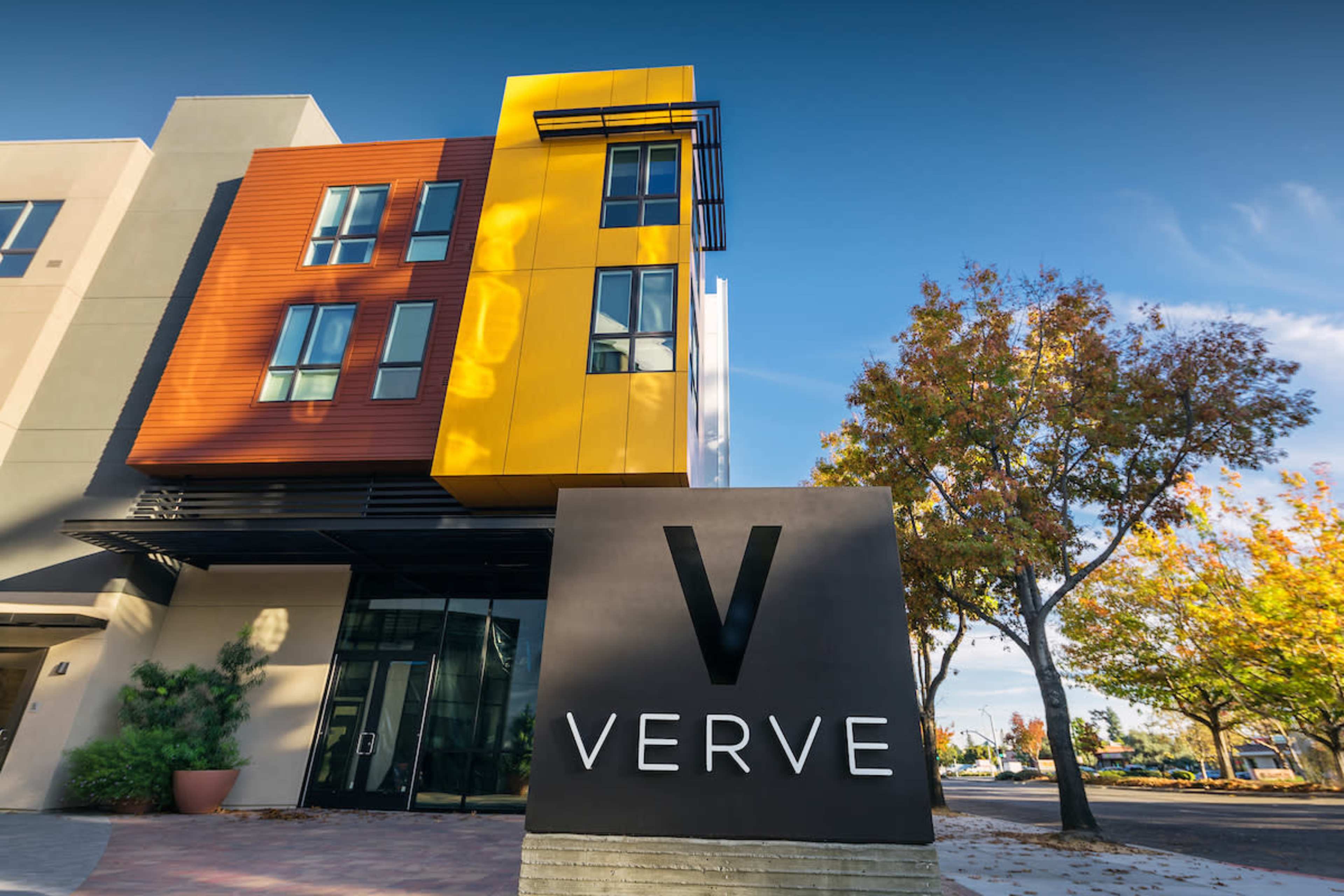 A modern building with a vibrant colored facade and a prominent sign displaying the word "VERVE" in front of it, surrounded by trees and clear blue skies.