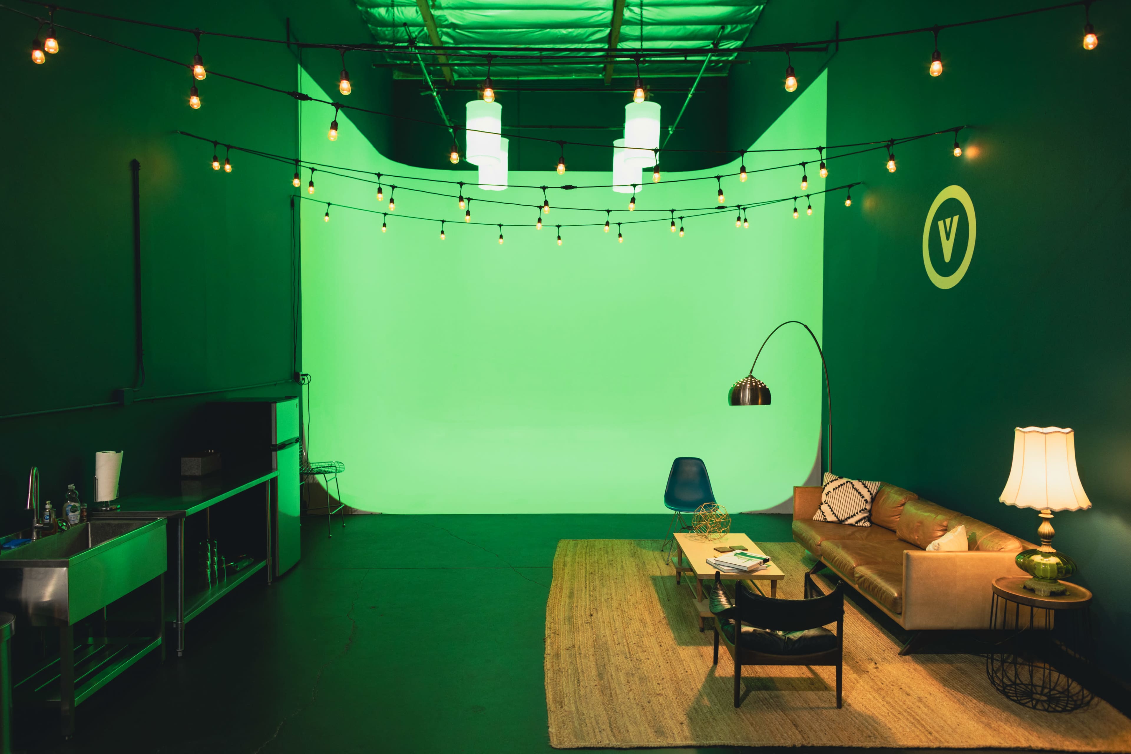 The image shows a green screen studio featuring a sofa, a coffee table, a floor lamp, and decorative string lights.