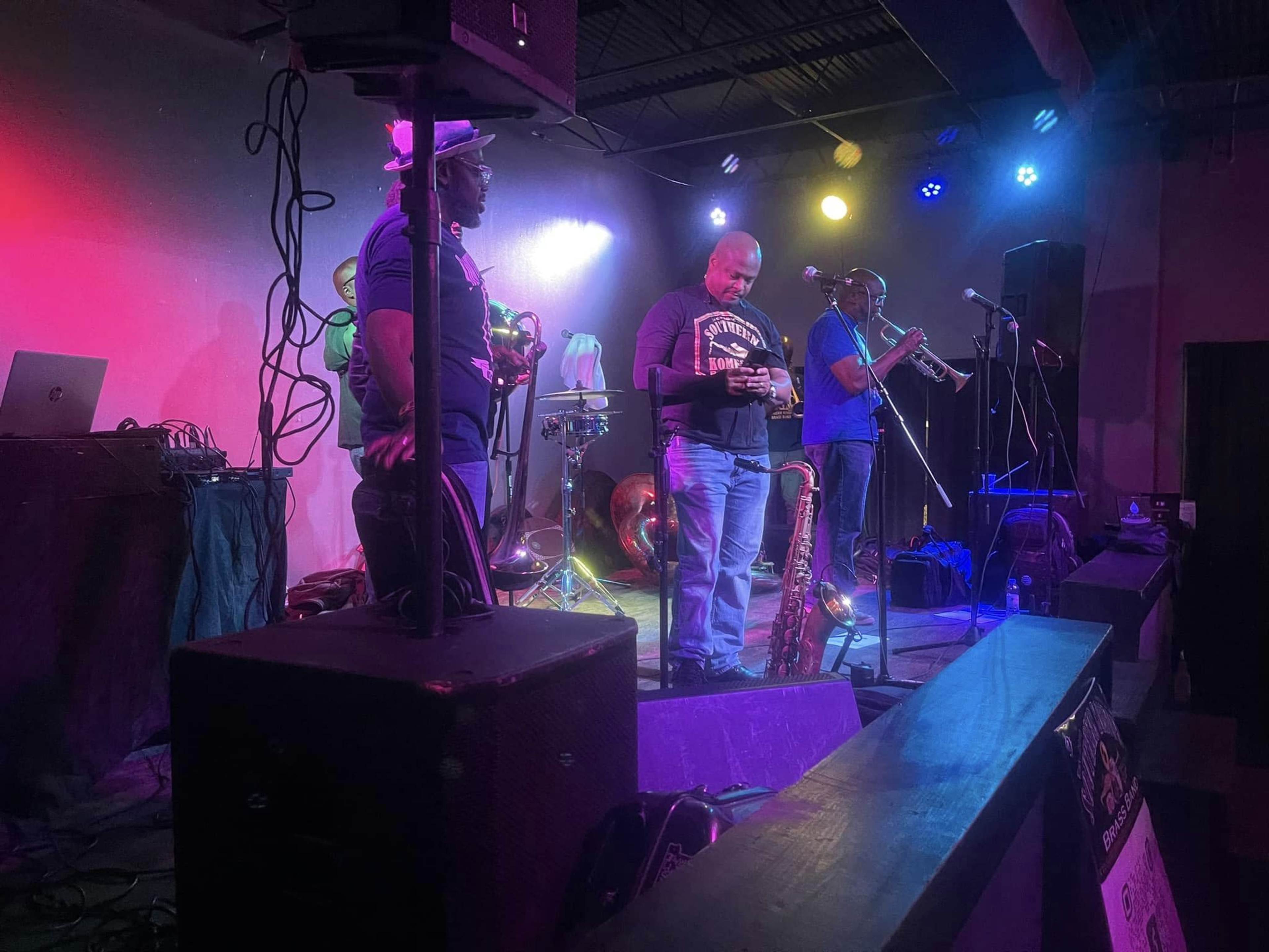 Three musicians stand on stage with brass instruments and microphones, preparing for a performance under colorful stage lights.