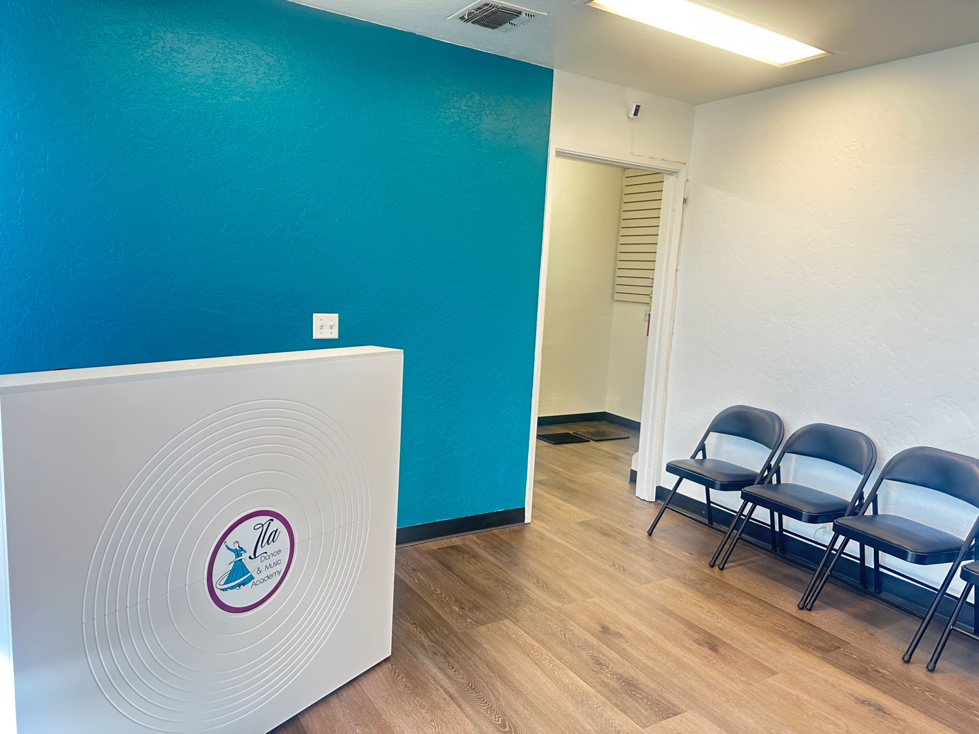 The image shows a reception area with a modern white desk and a teal accent wall, along with a waiting area featuring several black chairs.