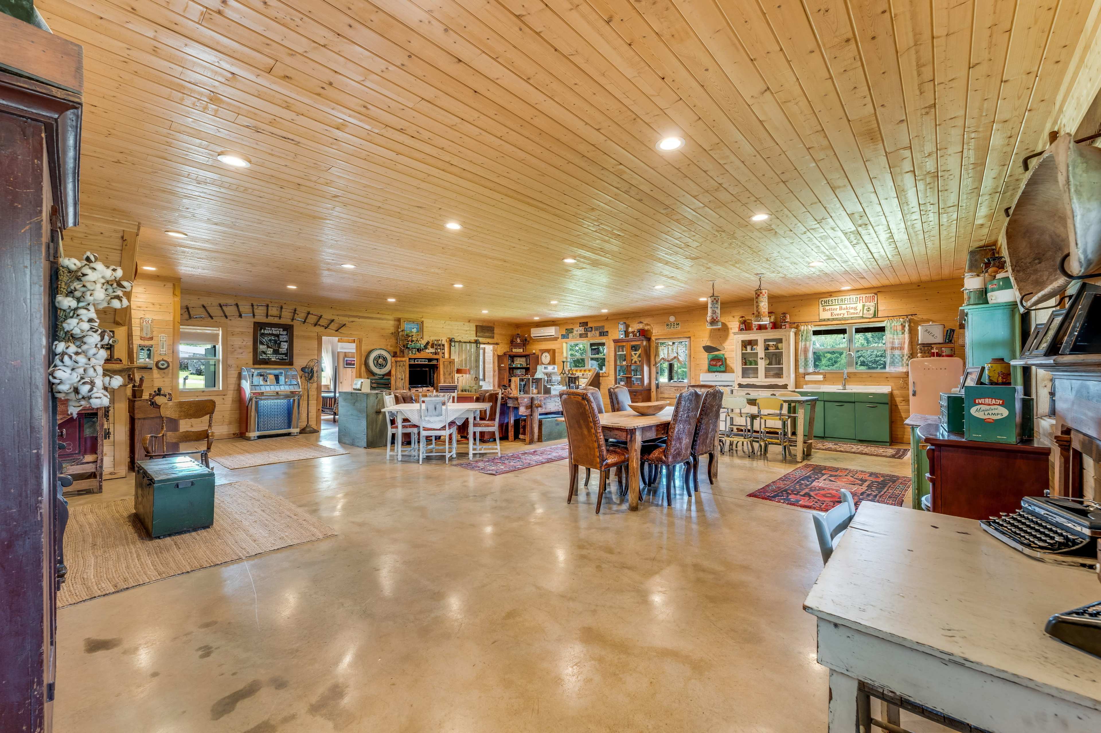 The image shows a spacious, well-lit room featuring a dining area with a large table, various furniture pieces, and rustic decor elements.