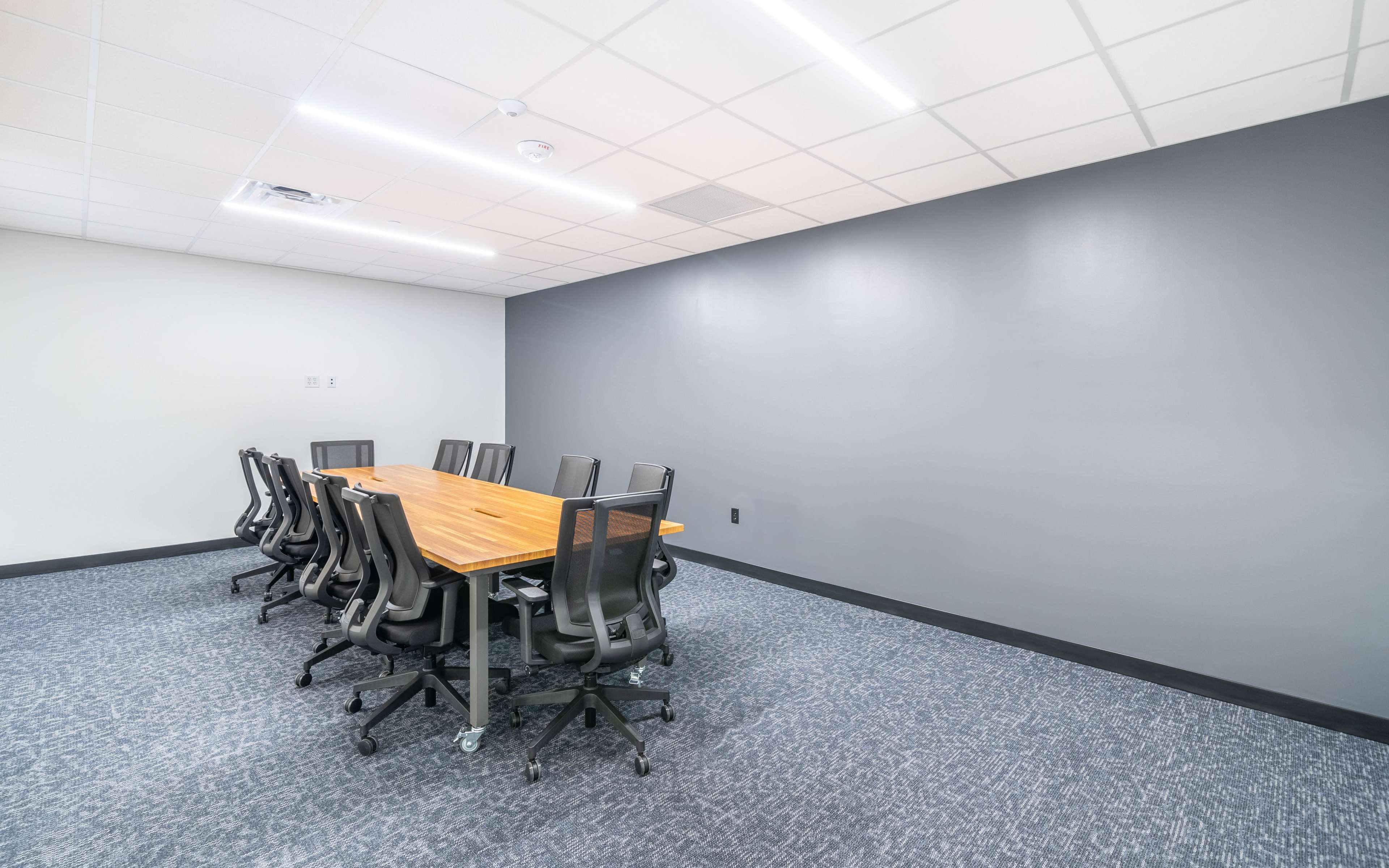 A long wooden conference table is surrounded by ten black office chairs in a minimalistic meeting room with gray walls and carpet.