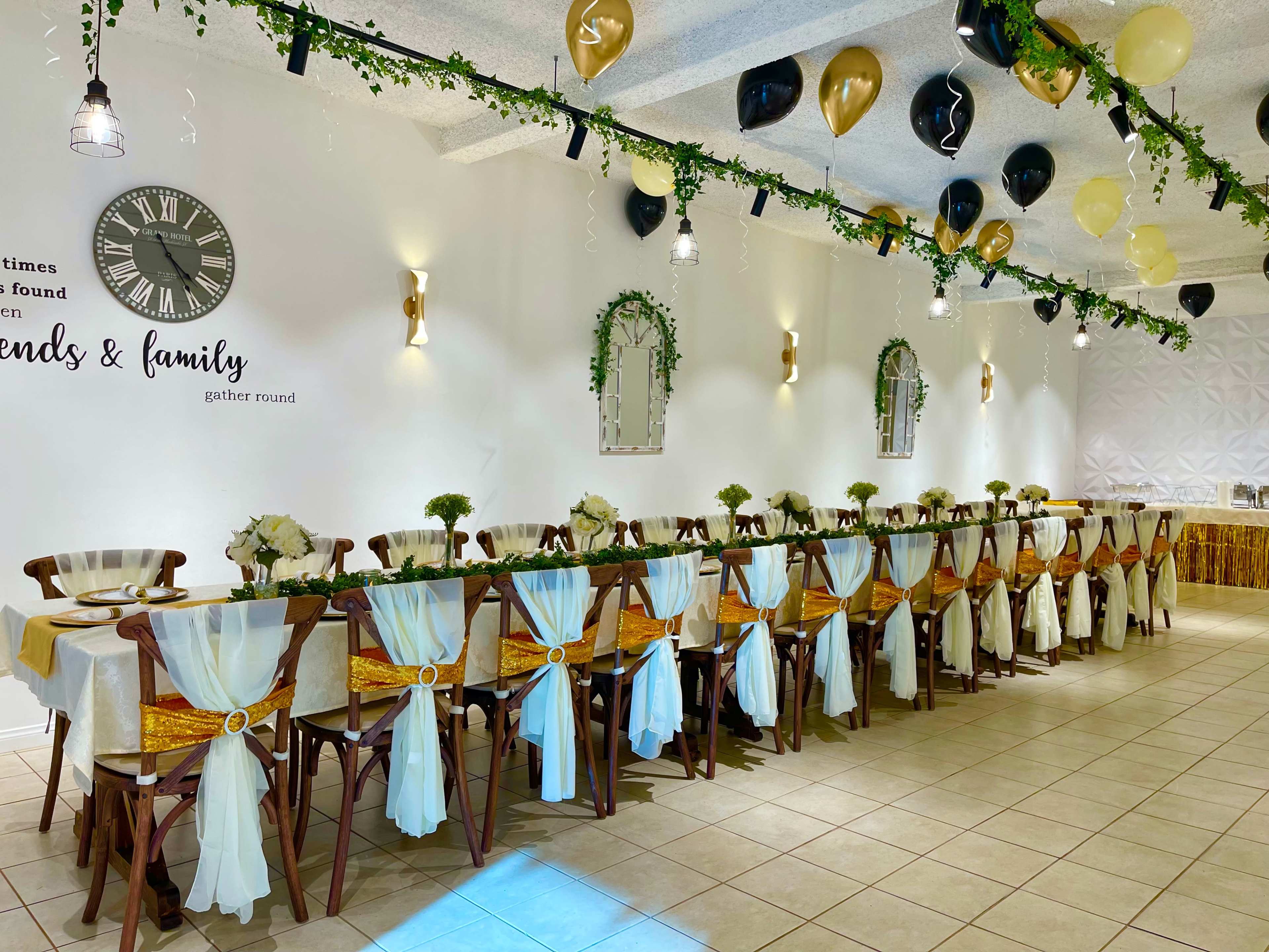 A long, elegantly set dining table is adorned with white and gold decorations, surrounded by wooden chairs and decorated with floral centerpieces, in a well-lit event space.