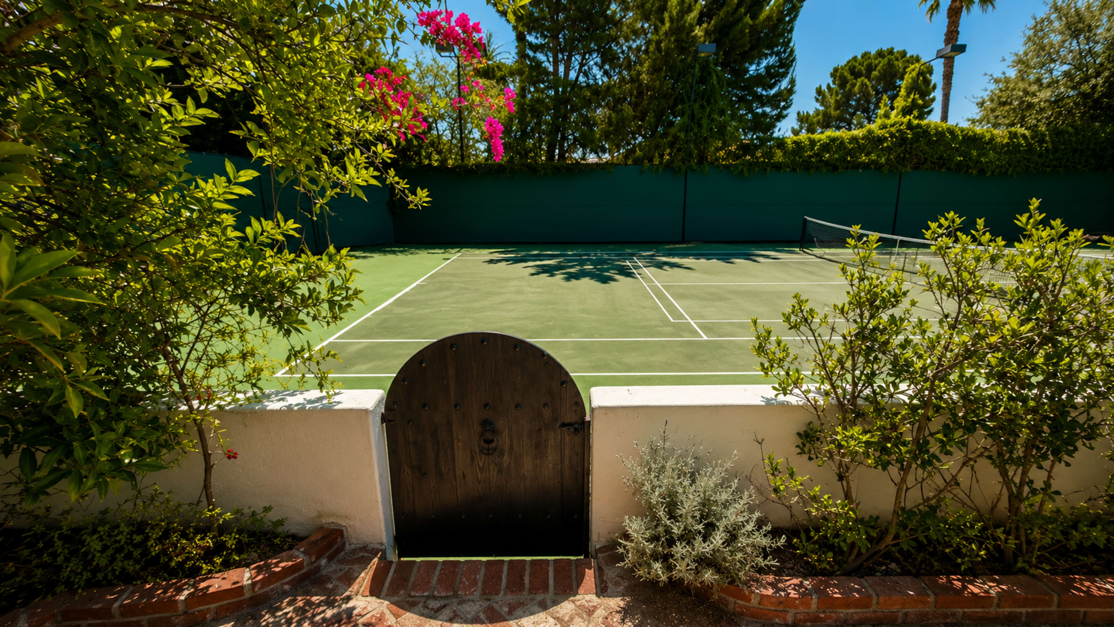 The image shows a well-maintained tennis court surrounded by lush greenery and flowers, with a small wooden gate in the foreground.