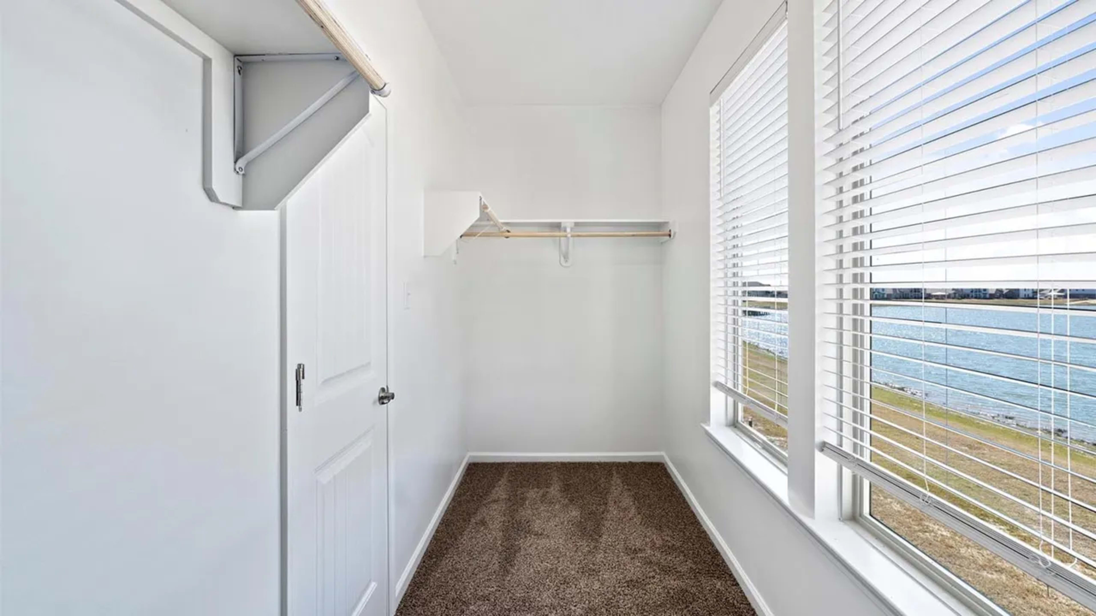 The image shows a small, empty room with a door, a hanging rack on the wall, and two windows with blinds overlooking a body of water.