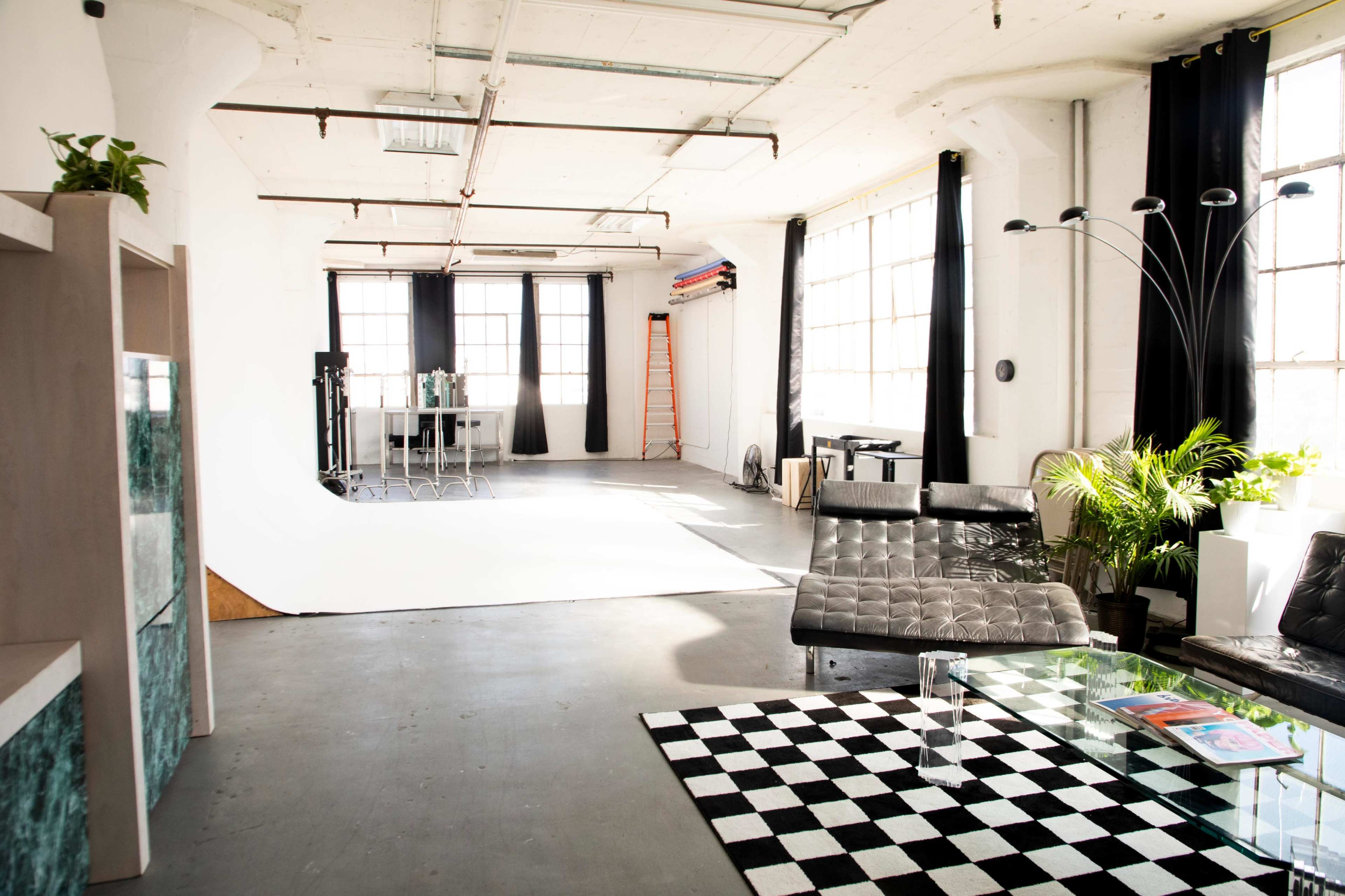 A spacious, well-lit studio featuring a checkerboard rug, a black sofa, a glass coffee table, and a background setup for photography.
