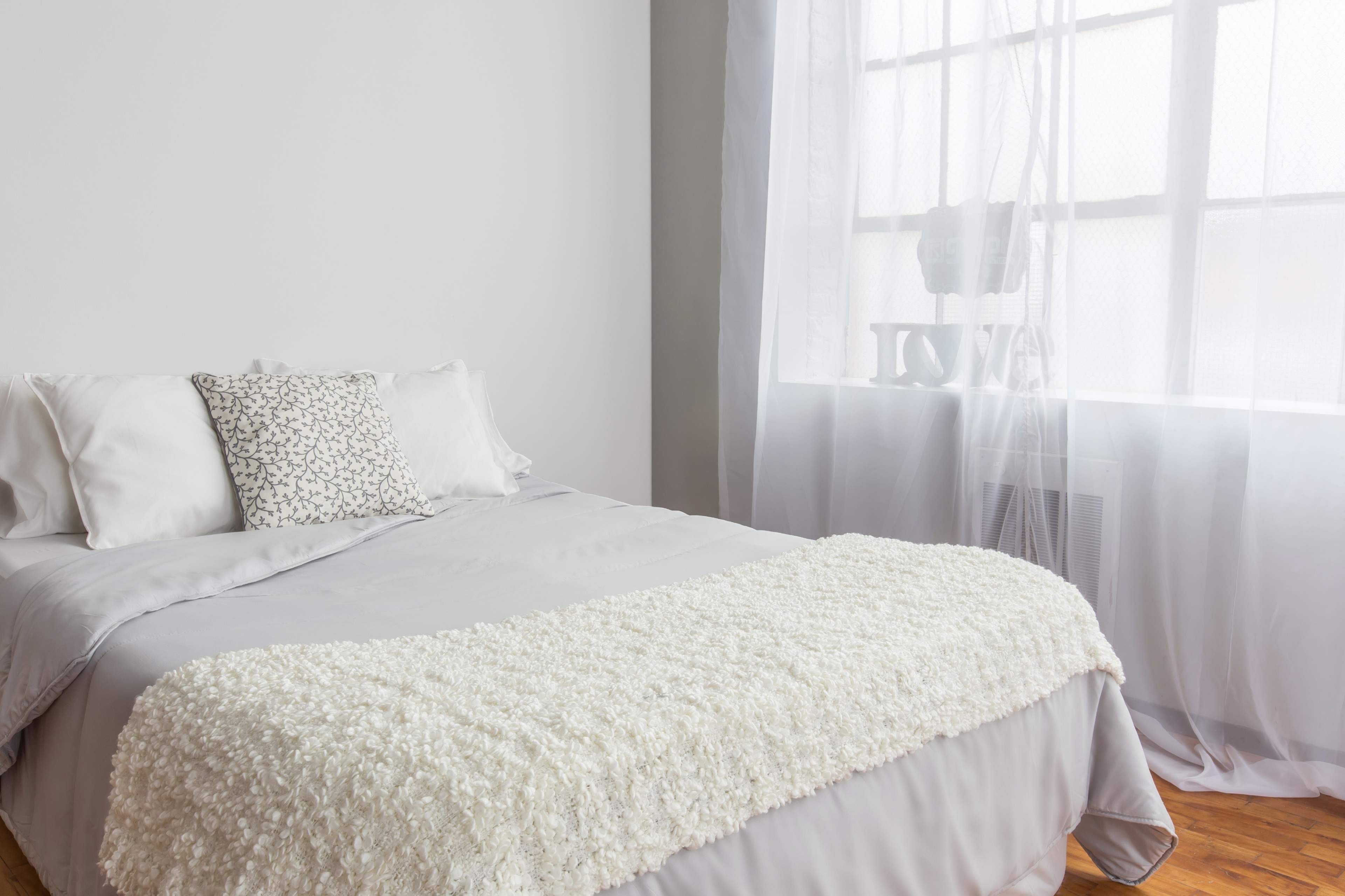 A neatly made bed with light gray bedding and decorative pillows is positioned beside a large window with sheer white curtains.