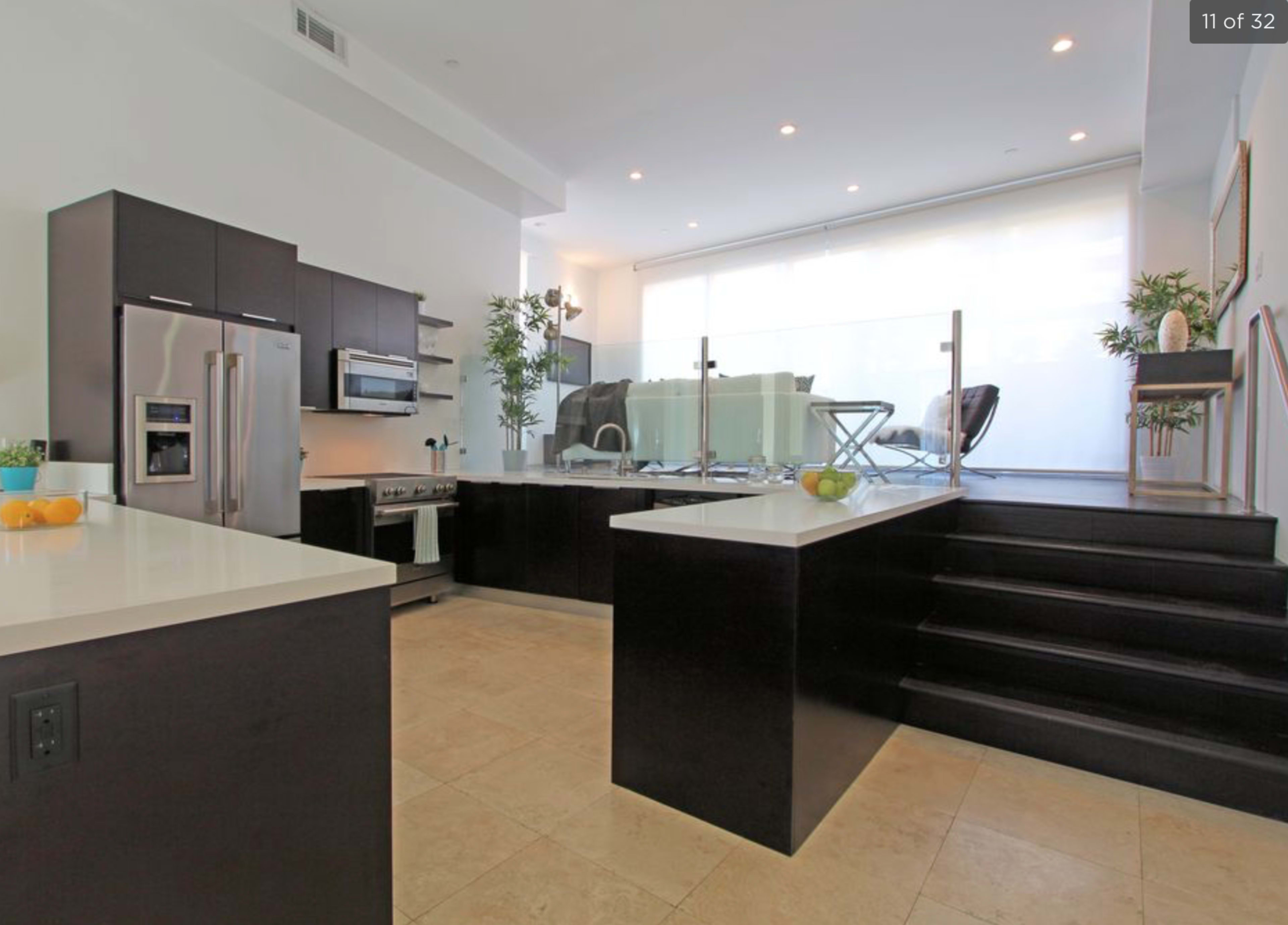 A modern kitchen with dark cabinetry, white countertops, and a glass partition leading to a living area with steps.