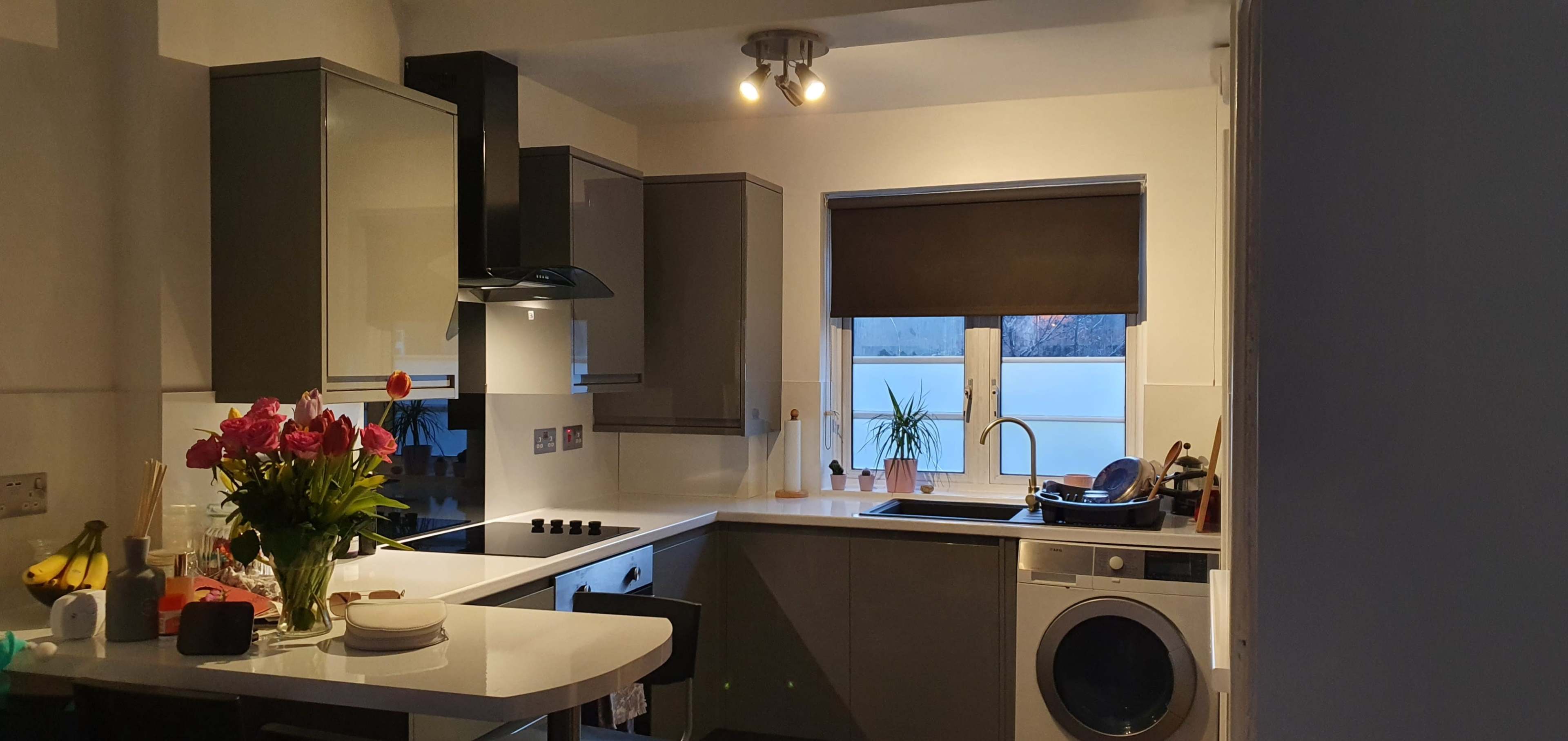 A modern kitchen with gray cabinets, a dark countertop, a window with a closed blind, and a washing machine beside the sink.