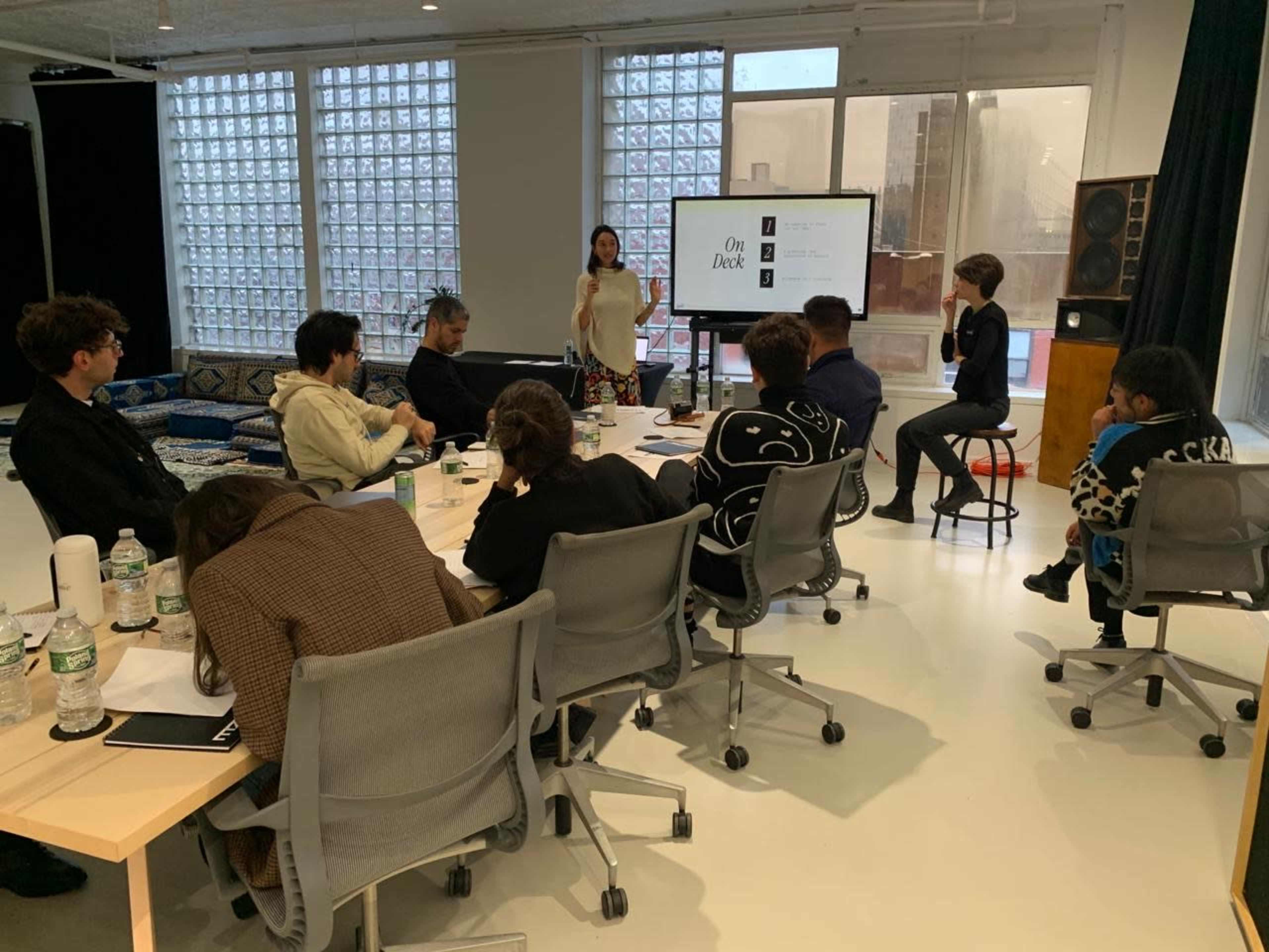 A group of people is seated around a table in a modern meeting room while a woman presents information on a screen.