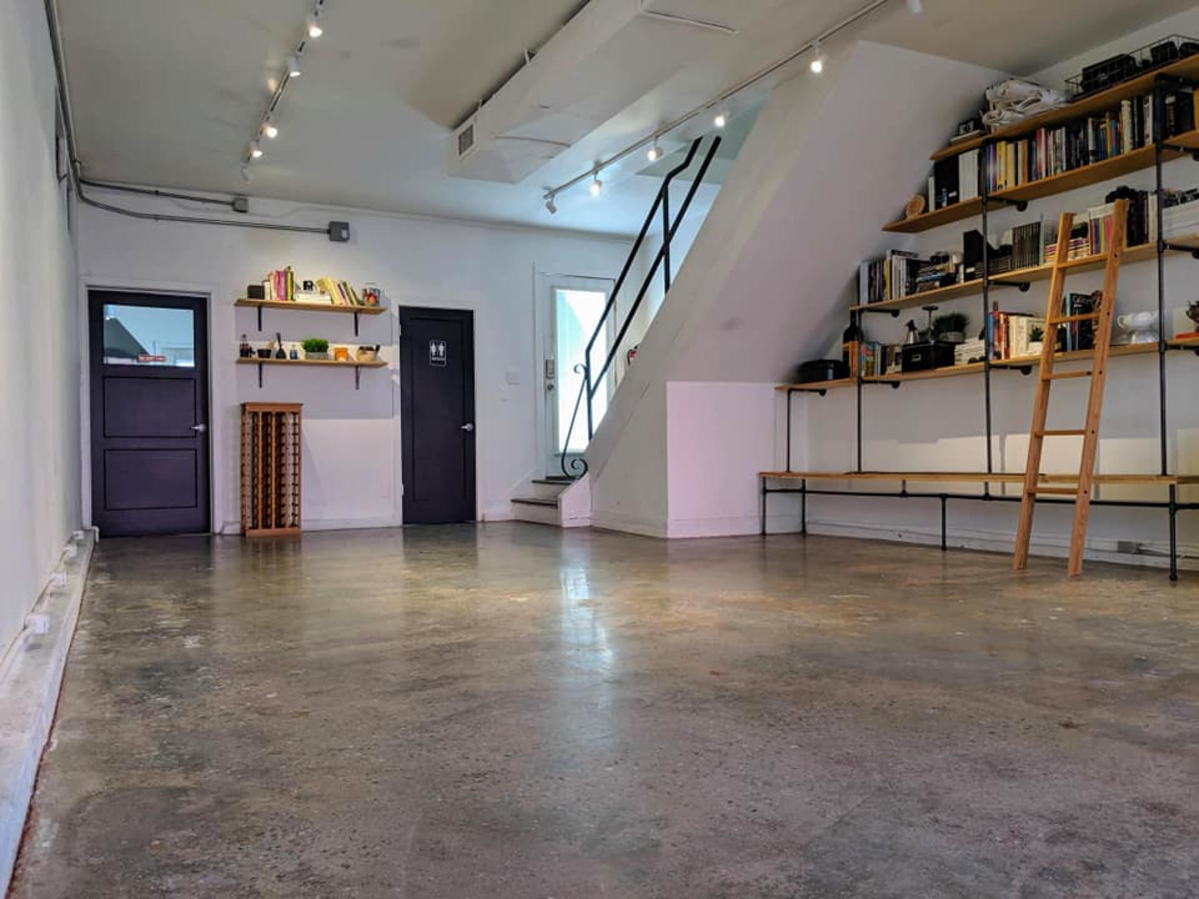 The image shows a minimalist interior space featuring a polished concrete floor, a staircase, shelves with books and plants, and a small entry area with doors.