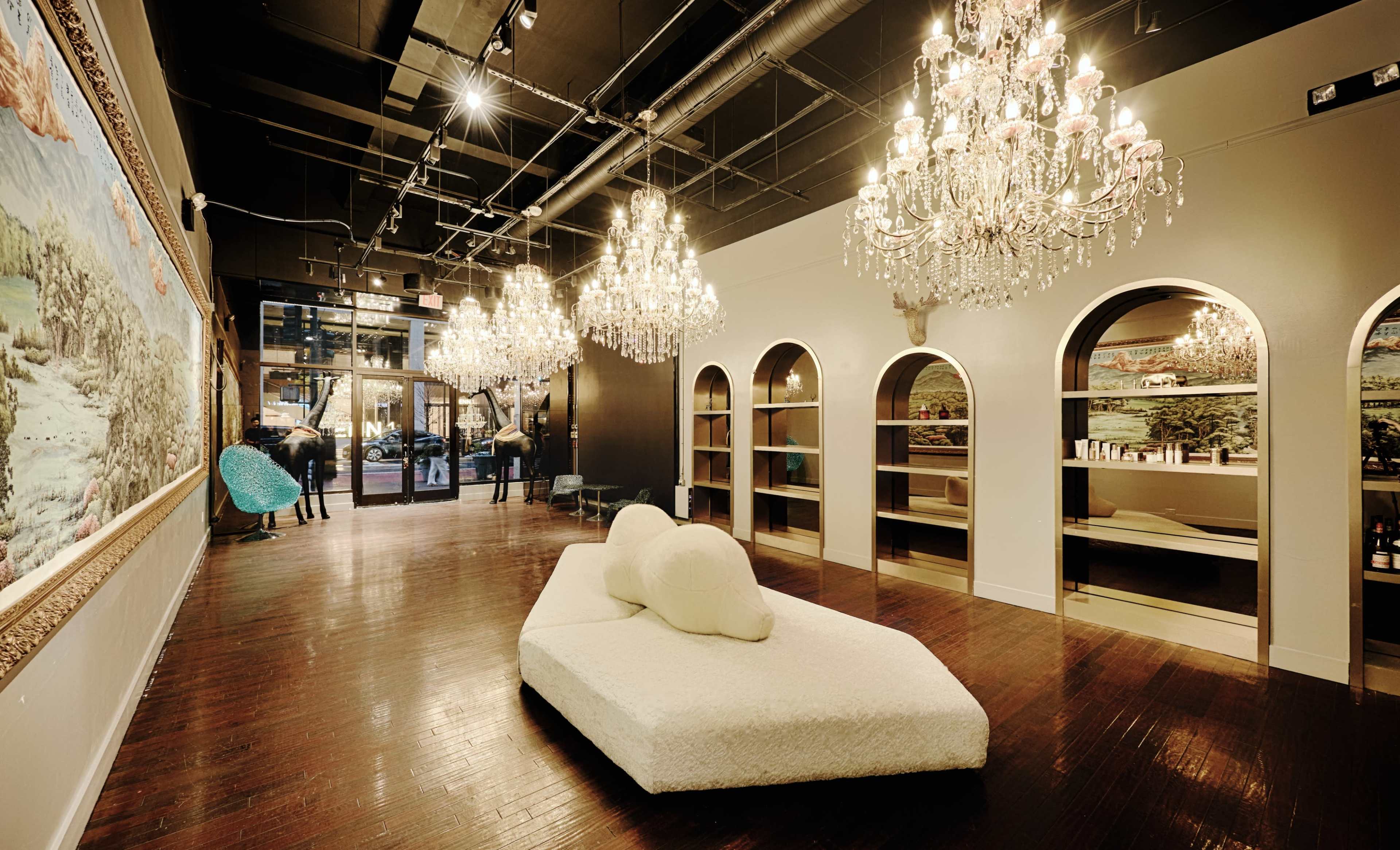 The image shows a spacious, modern interior with multiple chandeliers, a white sculptural sofa, and arched shelving on the walls.