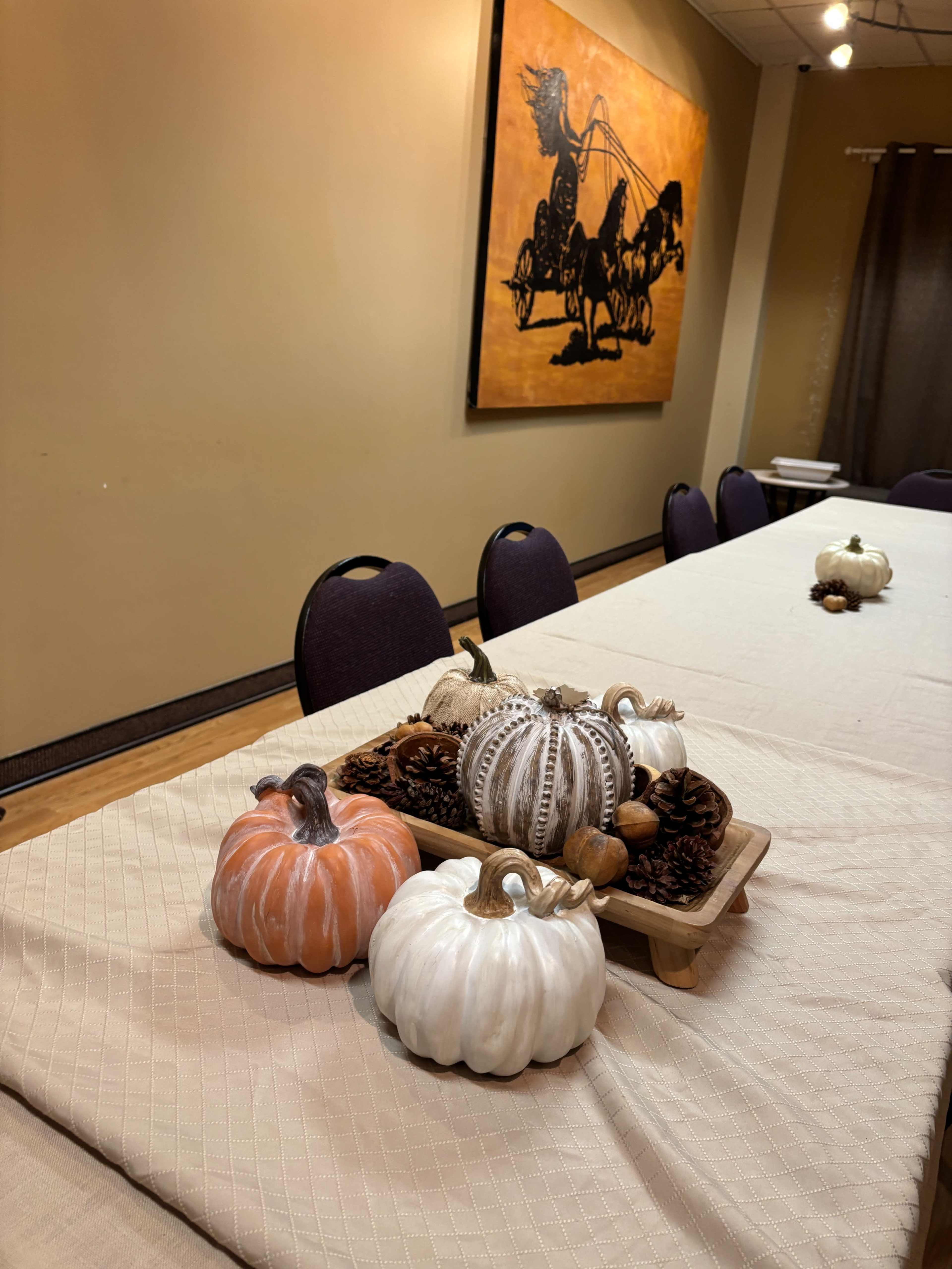 A long table is set with a beige tablecloth and decorated with decorative pumpkins and pine cones at one end, against a background featuring a large painting of a horse-drawn carriage.