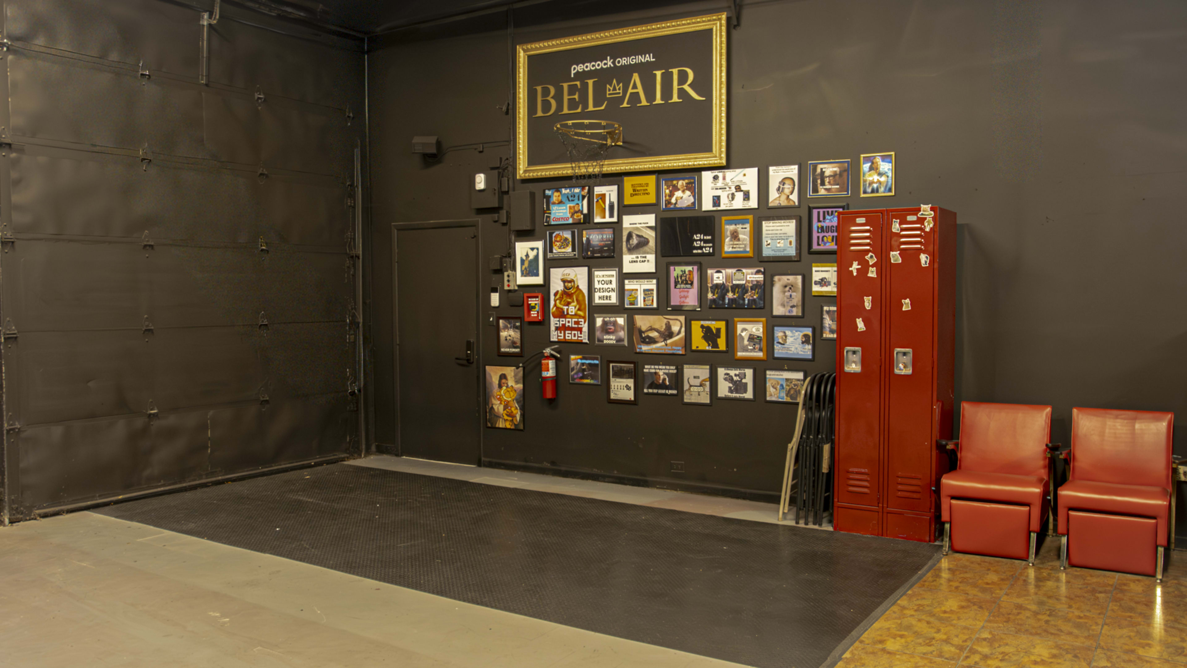 The image shows a blank stage area with a black wall featuring a collage of framed pictures, a red locker, and two red chairs.