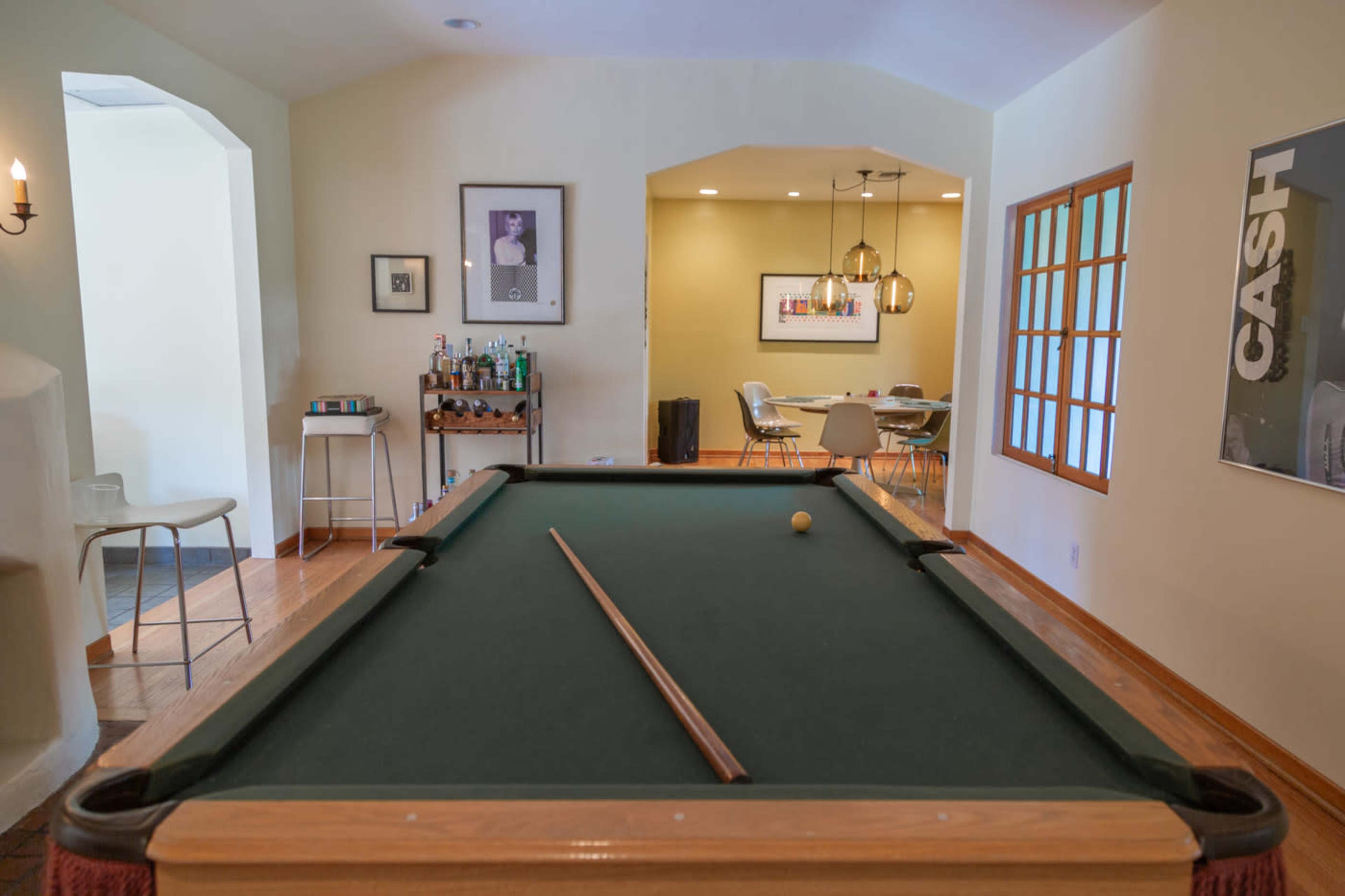 A pool table in the foreground with a cue stick and a single ball, leading to a well-lit dining area in the background that features a table and chairs.