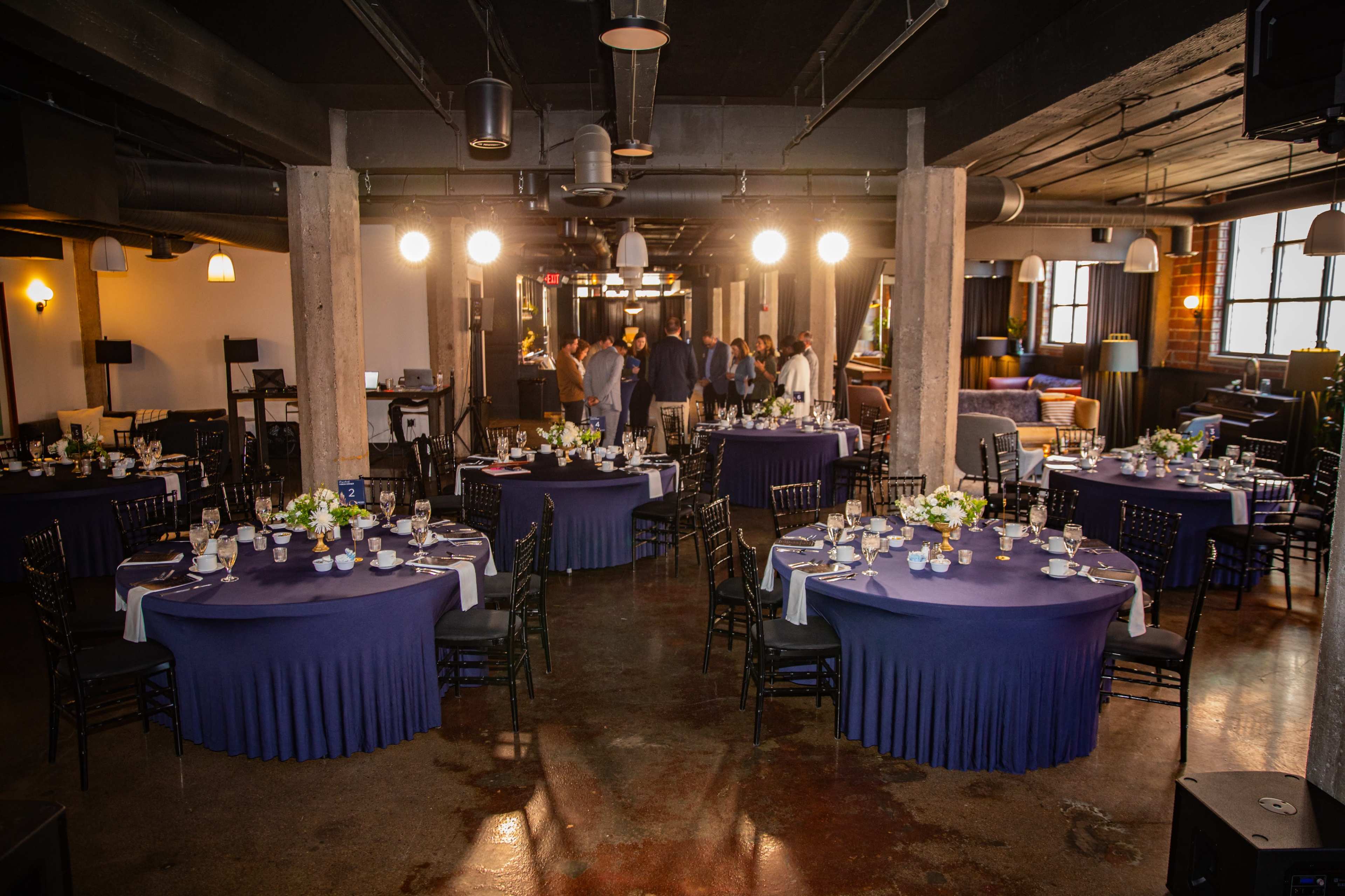 A dining area is set up with several round tables covered in dark blue linens, surrounded by chairs, while guests gather in a group in the background.