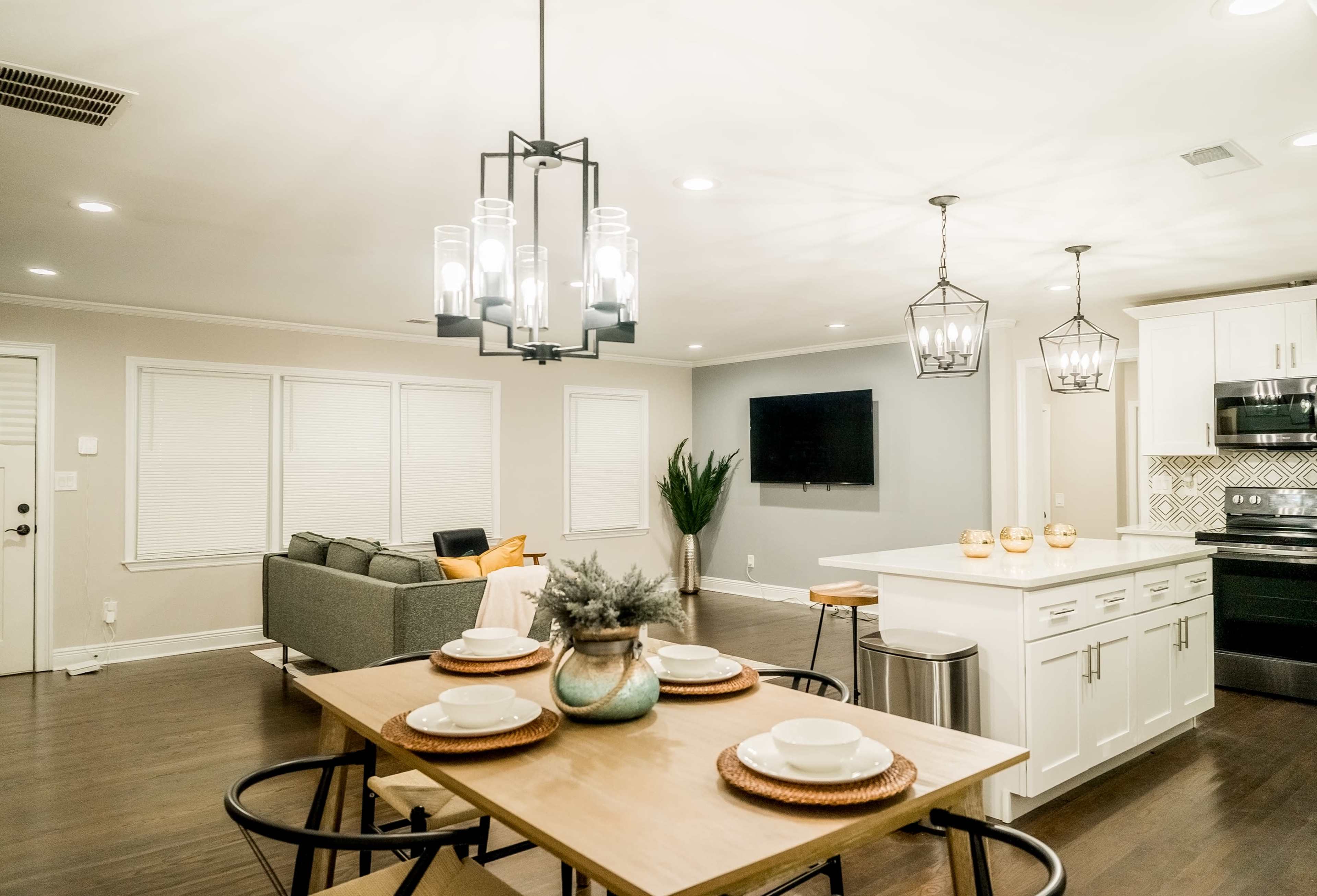 A modern open-concept living area features a dining table set for four, a well-lit kitchen island, and a cozy seating area with a gray sofa.
