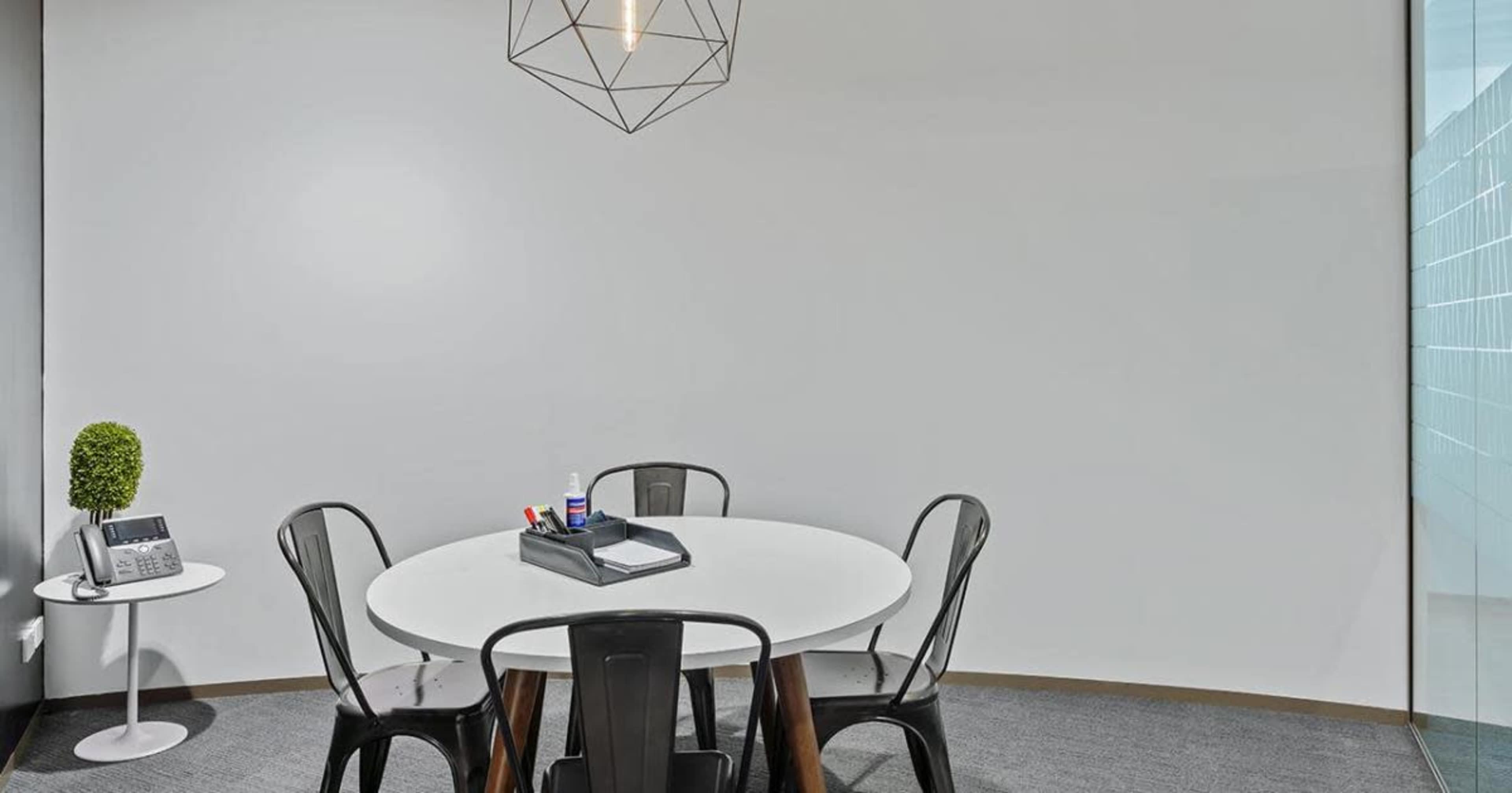 A round table surrounded by black chairs is set in a minimalist office space with a small plant and office supplies on the table.