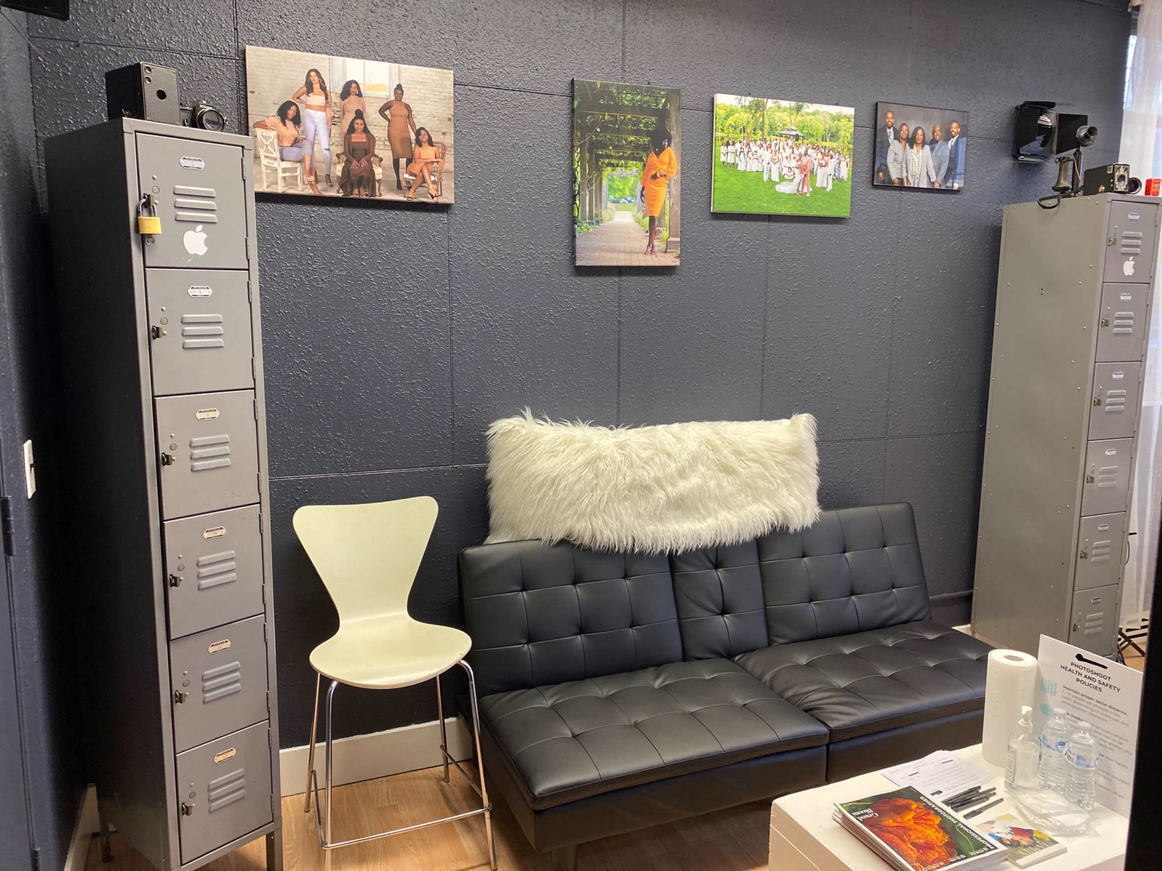 A modern sitting area with a black couch, a white chair, gray lockers, and framed photographs on a dark wall.