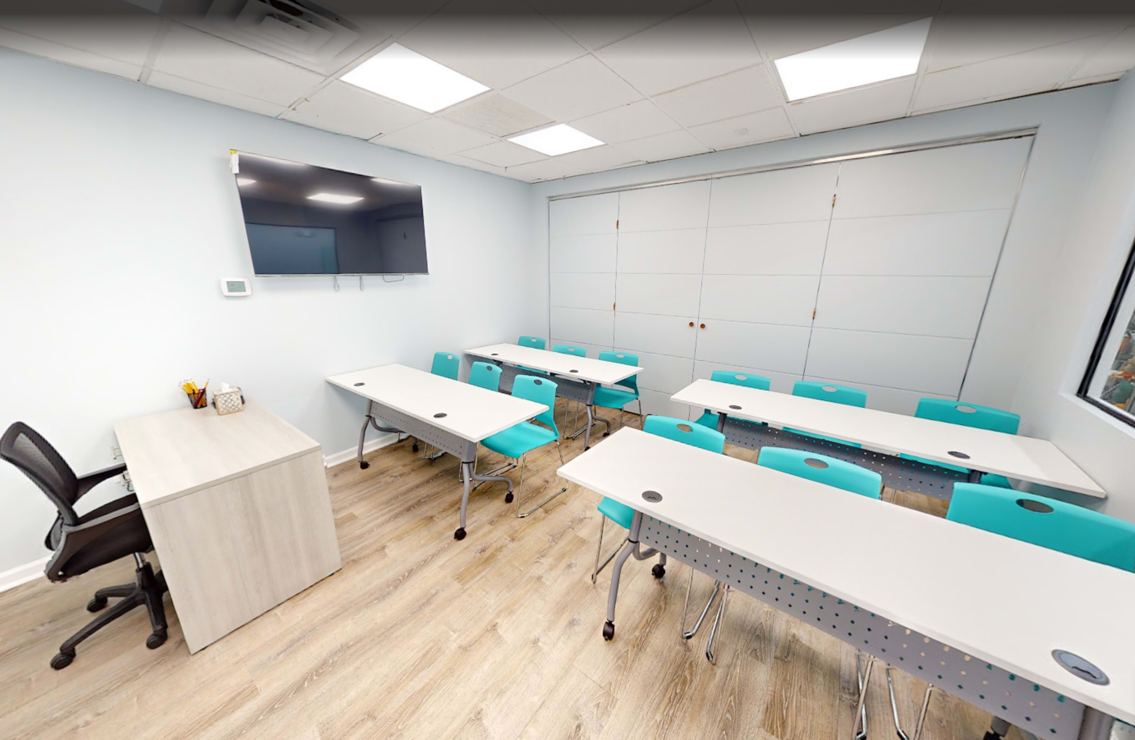 The image shows a modern classroom with several tables and chairs arranged in a U-shape around a central workspace, along with a television mounted on the wall.