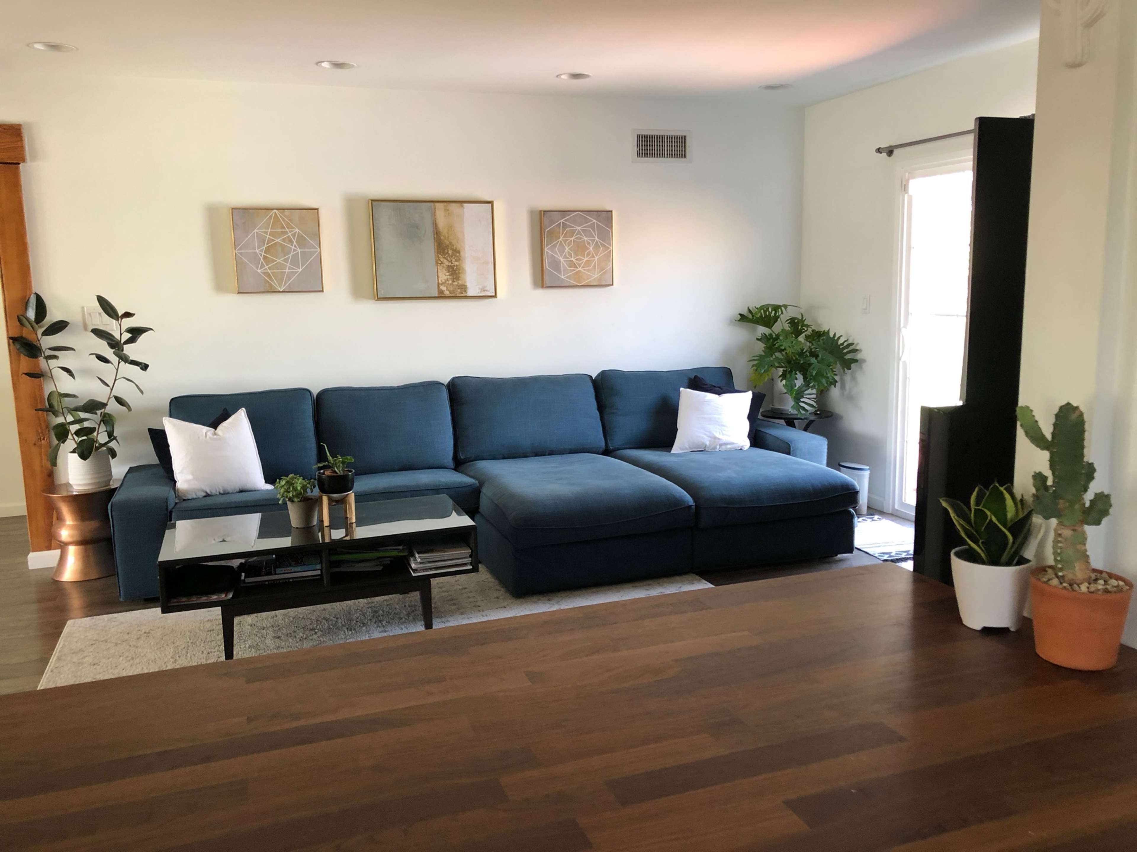 A large blue sectional sofa sits in a well-lit living room, accompanied by plants and decorative wall art.