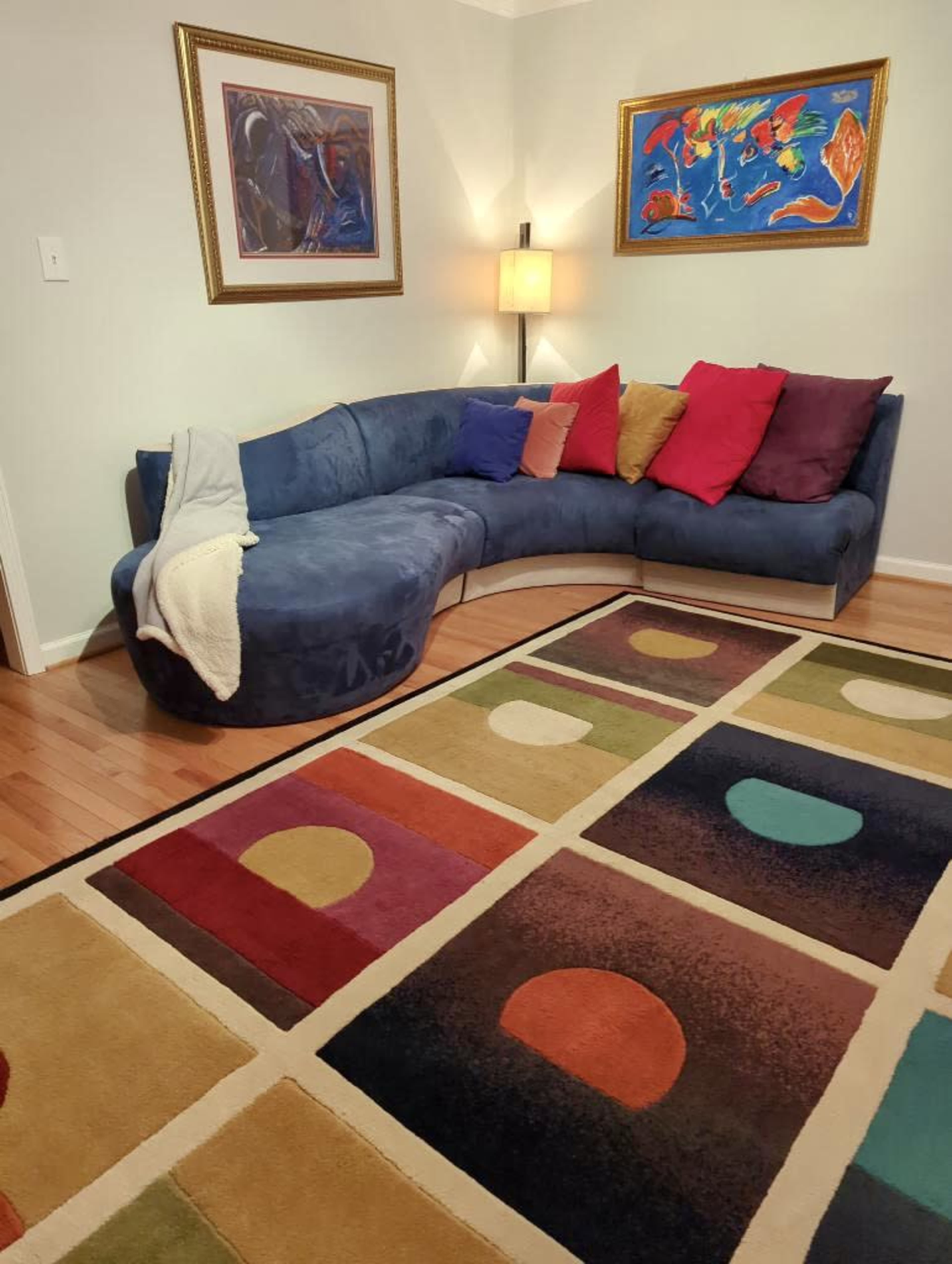 A living room with a curved blue sofa adorned with colorful pillows, a floor lamp, and a patterned area rug featuring geometric shapes.