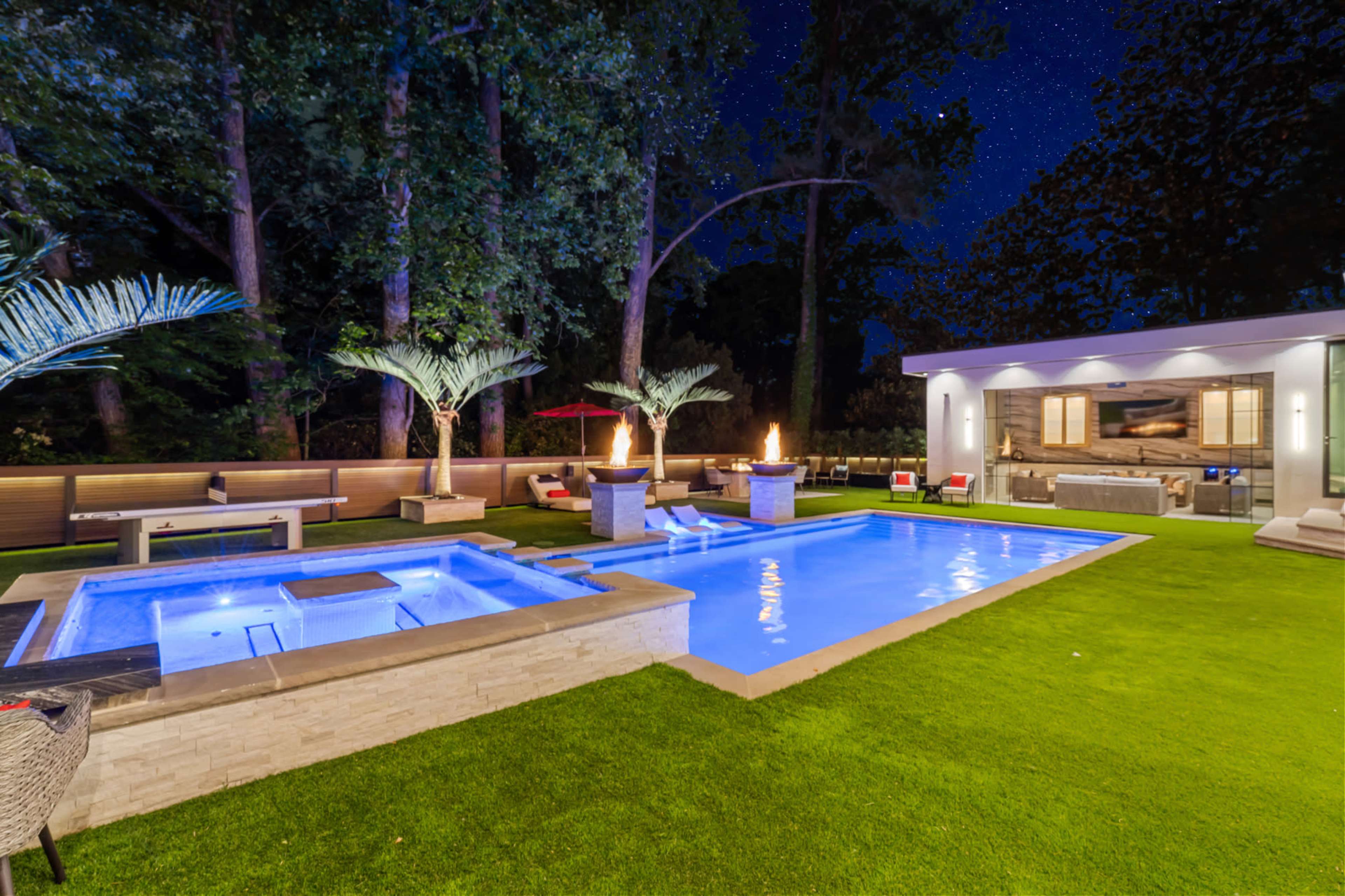 The image shows a modern outdoor space at night, featuring a lit swimming pool, a hot tub, and landscaped greenery with fire features and seating areas.