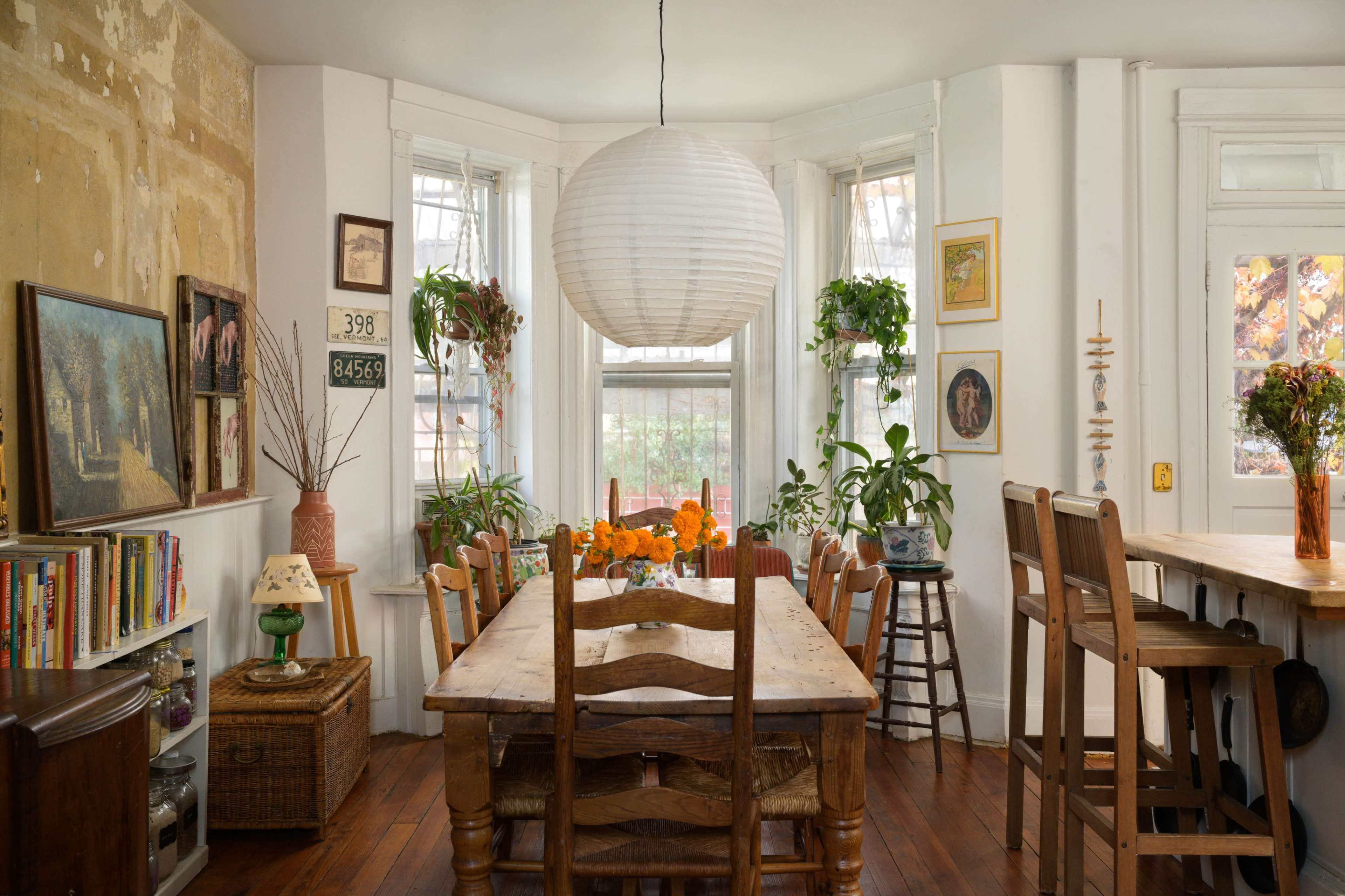 A spacious dining area features a long wooden table surrounded by chairs, illuminated by a round paper lantern, with plants and artwork adorning the walls.