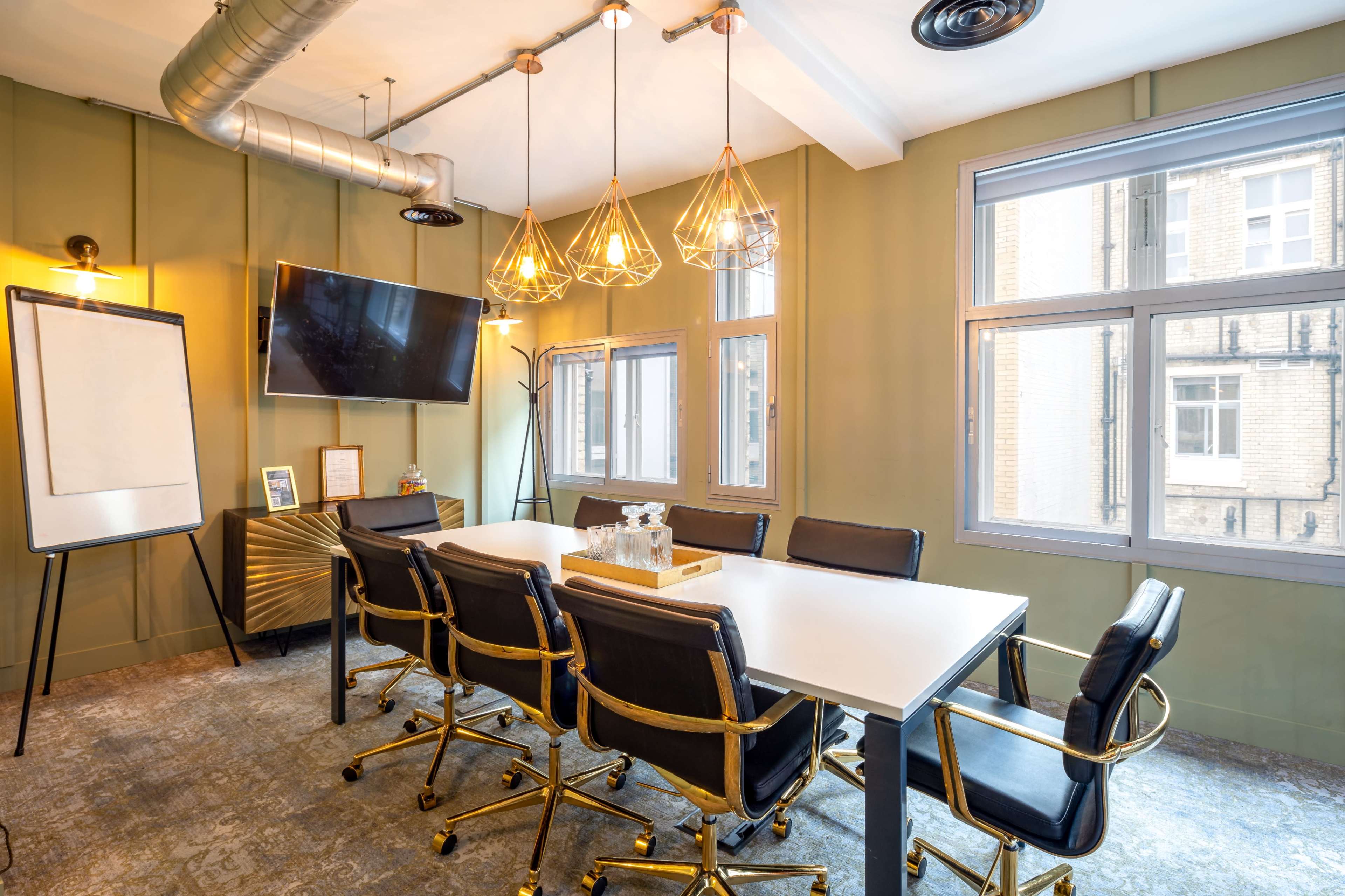 A modern conference room features a long table surrounded by black and gold chairs, a television mounted on the wall, and three pendant lights above.