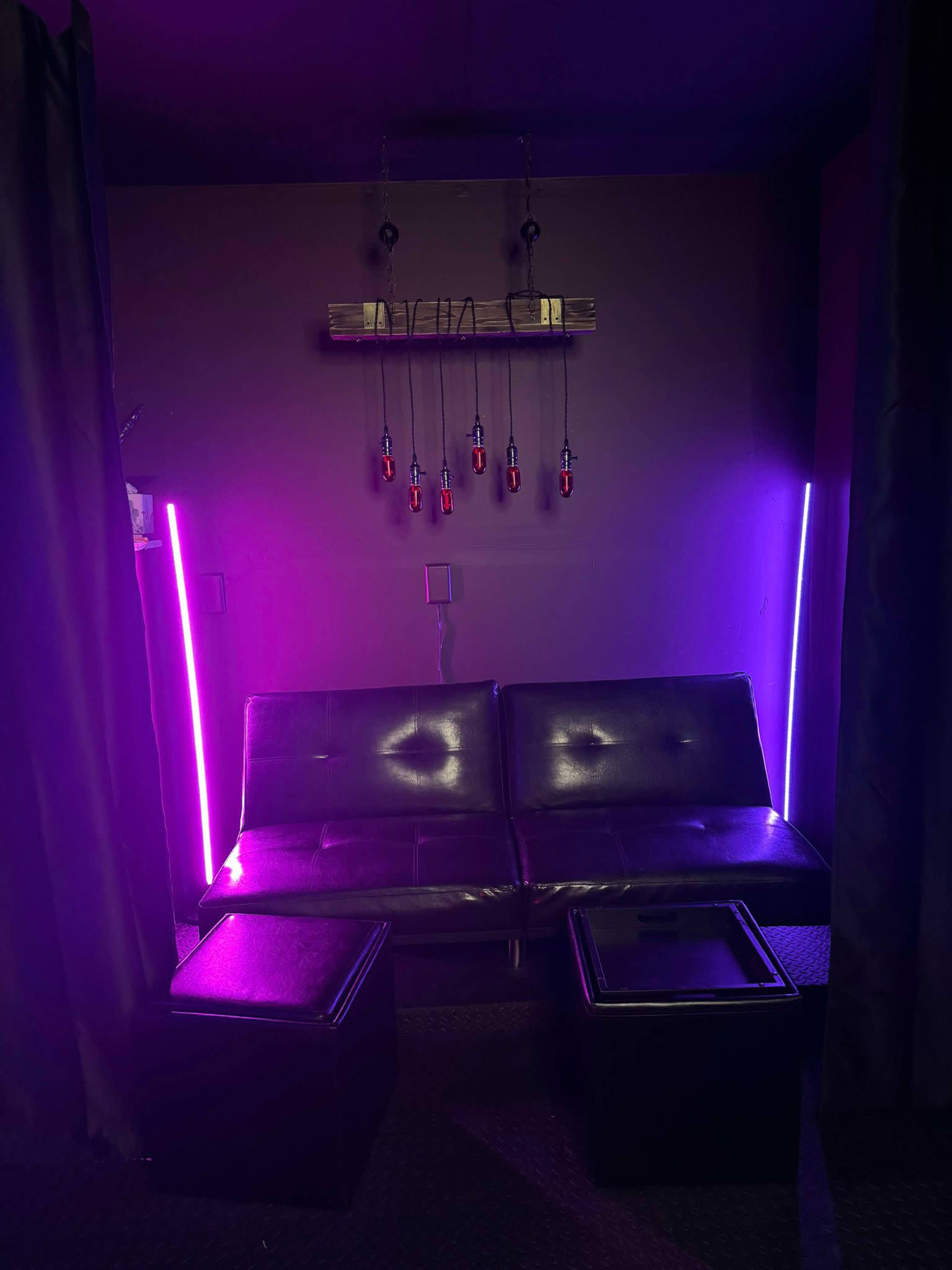 The image shows a small lounge area with a dark sofa, two black stools, and purple and pink LED lights illuminating the space.