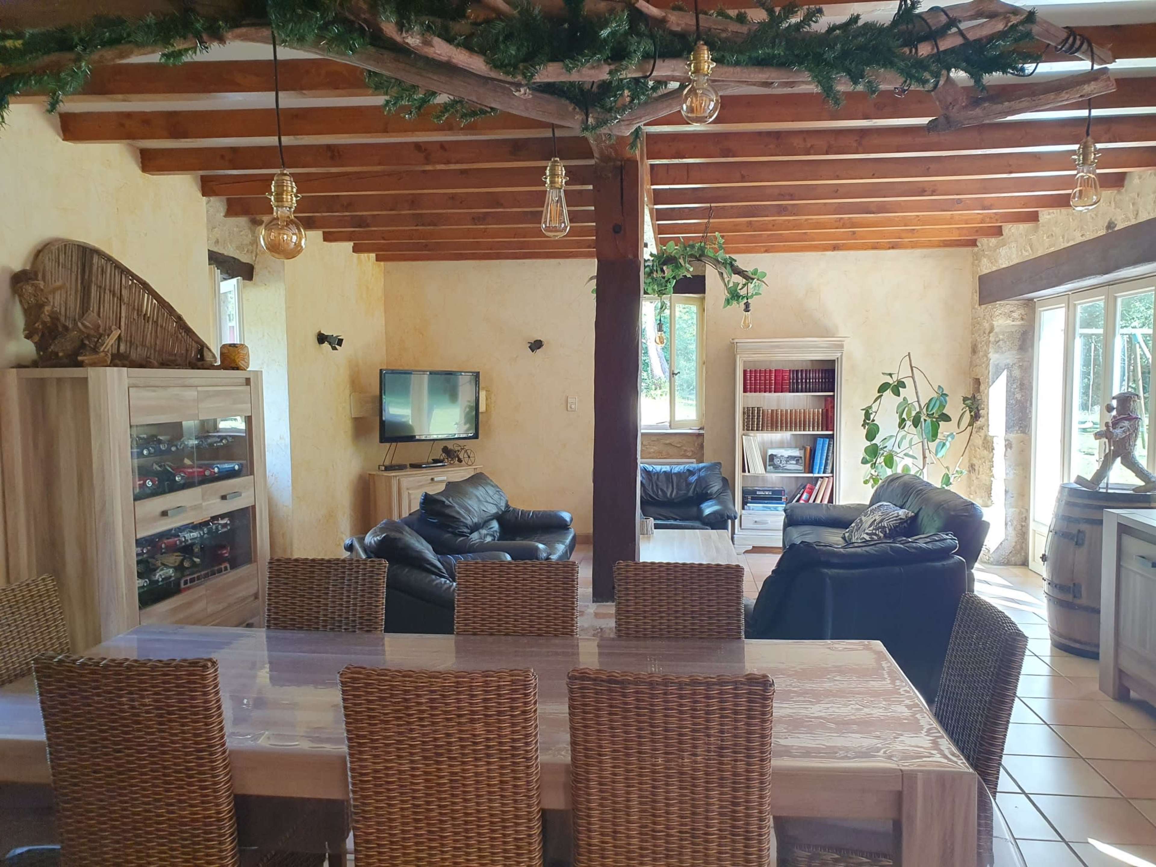 The image depicts a cozy interior of a living and dining area featuring a wooden ceiling, a glass-top dining table with wicker chairs, and comfortable black leather seating.