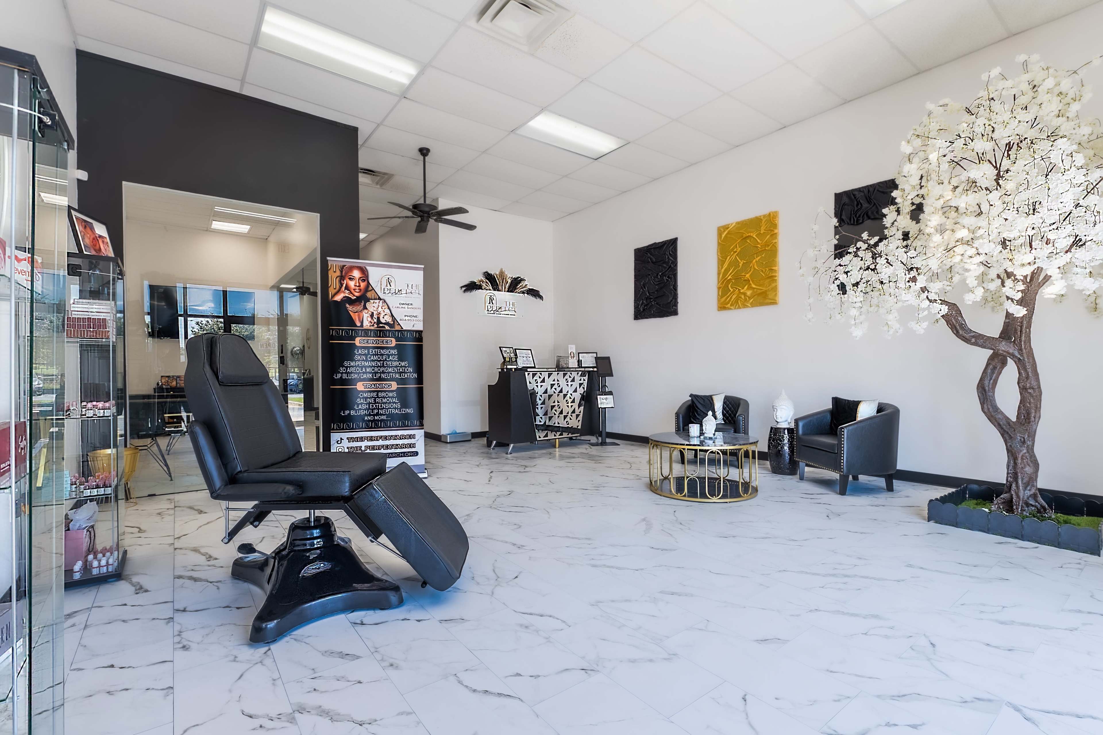 A modern salon interior with marble flooring, a black styling chair, decorative wall art, and a faux flowering tree in a corner.