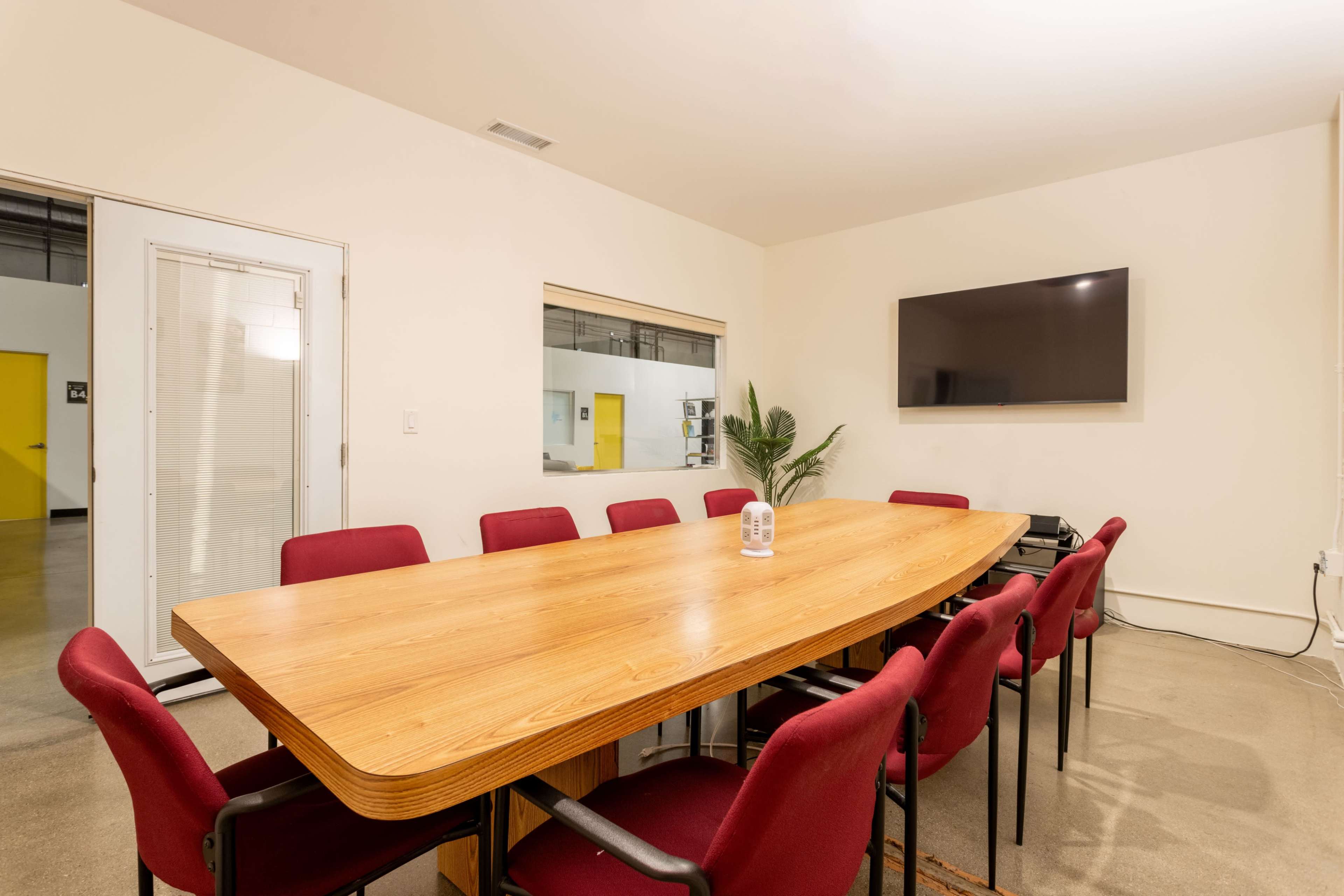 A long wooden conference table is surrounded by maroon chairs in a meeting room featuring a television and a plant.