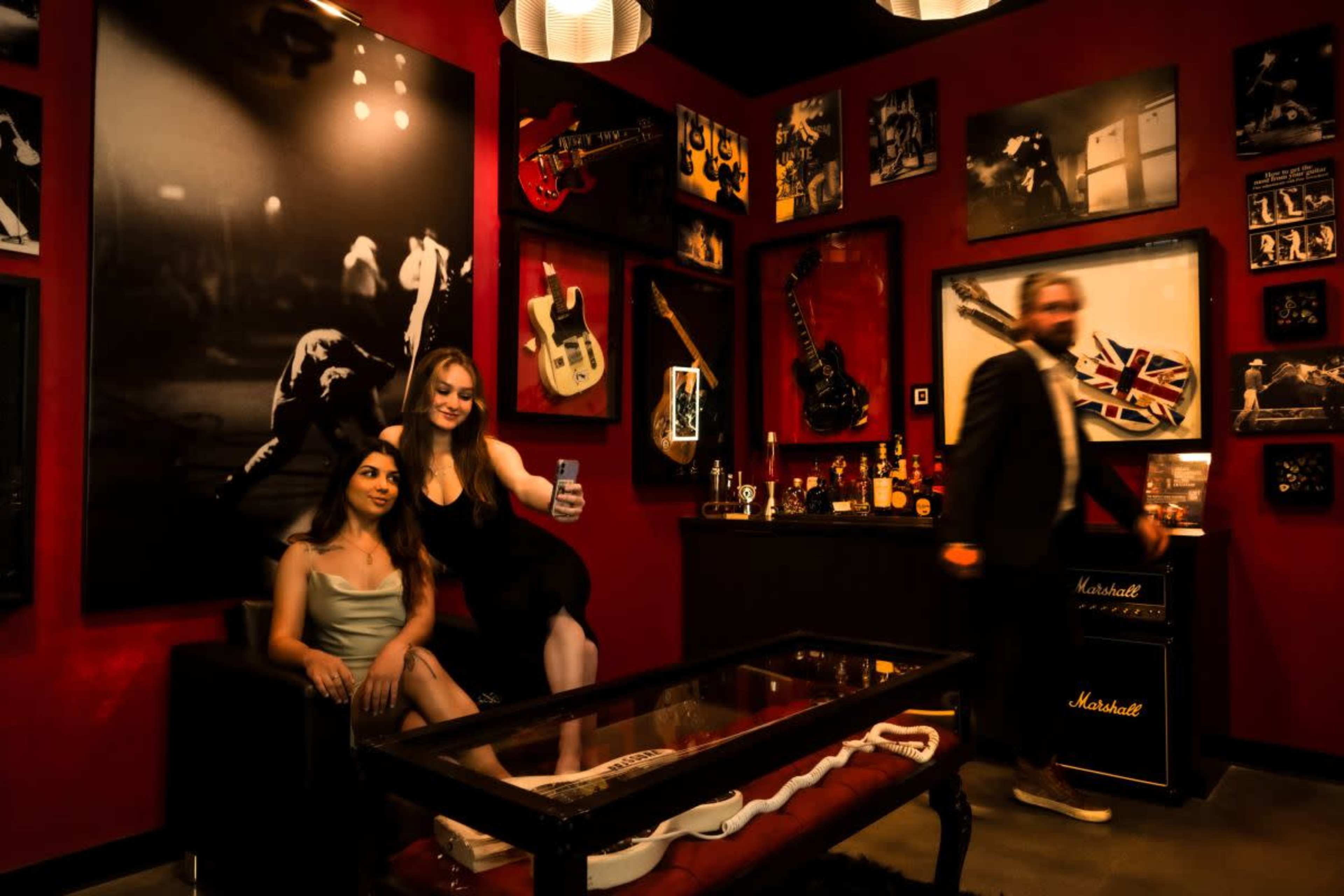 Two women sit on a couch in a room decorated with guitars and framed music photographs, while a man walks by in the background.